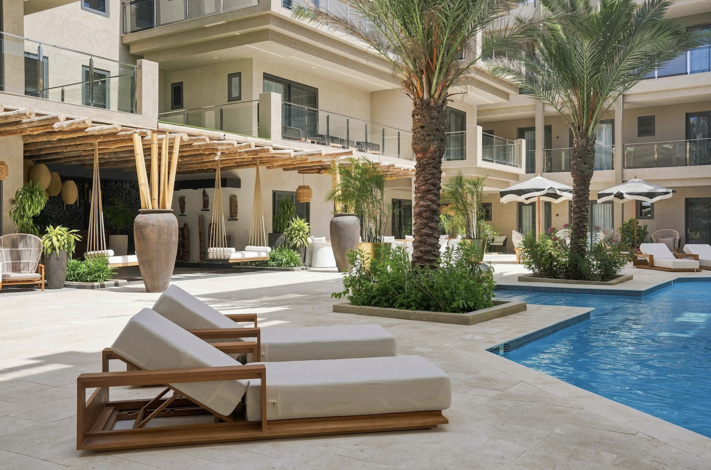 A luxurious hotel pool area with lounge chairs, palm trees, and umbrellas, surrounded by modern building balconies.
