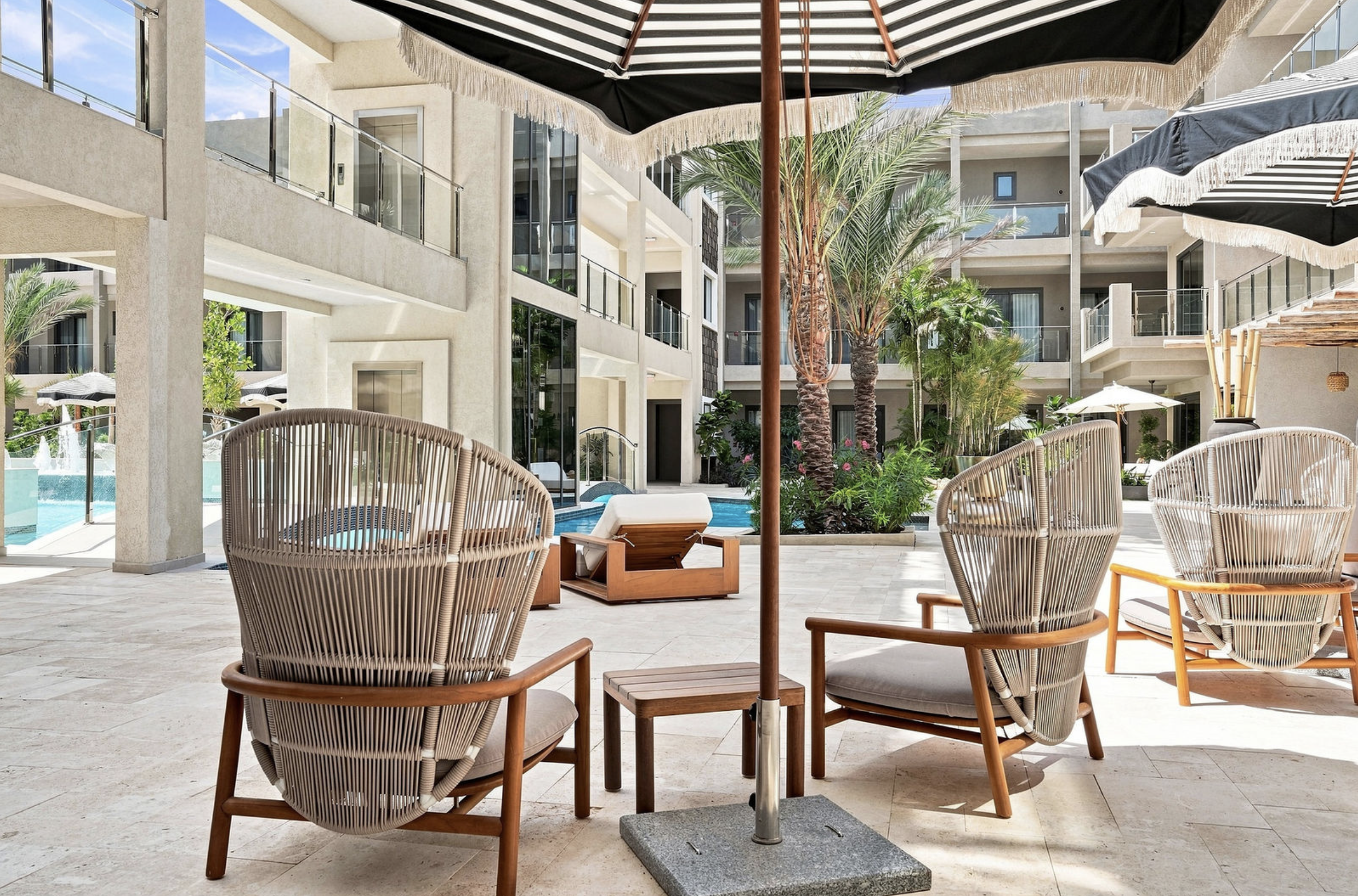 Poolside lounge area with wicker chairs, umbrellas, and greenery at a modern hotel courtyard.