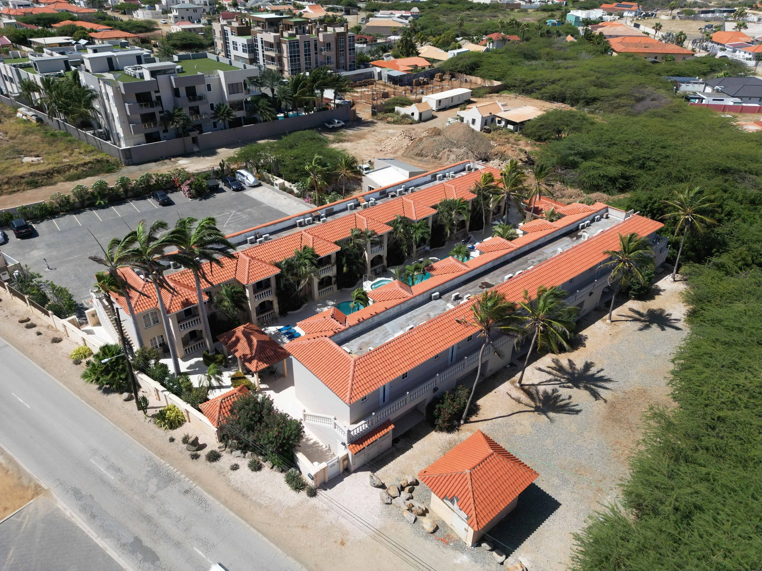 Aerial view of a residential complex with red-tiled roofs, palm trees, and a swimming pool, surrounded by parking spaces and other buildings.