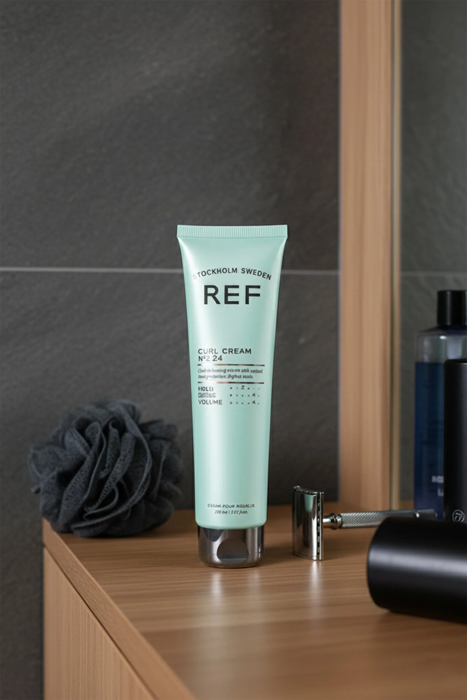 A tube of REF Curl Cream on a wooden surface in a bathroom. To the left is a black scrunchie, and to the right are a safety razor and a black bottle.