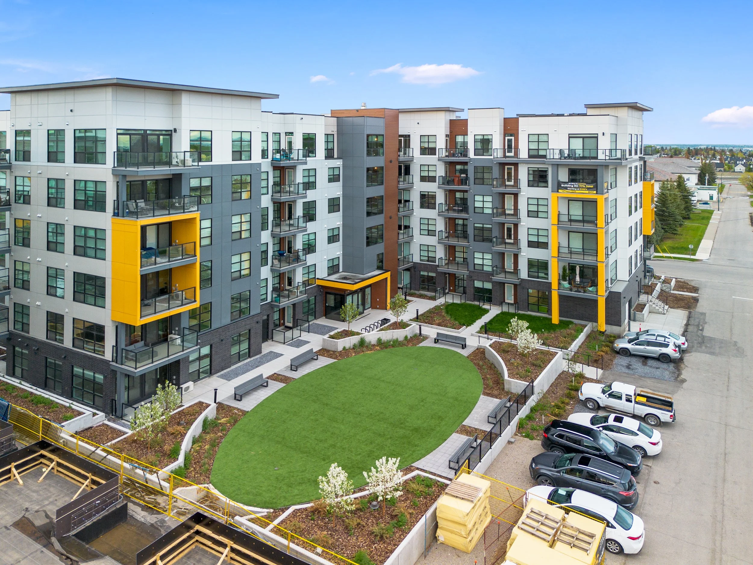 Modern multi-story apartment complex with colorful yellow accents surrounding a landscaped courtyard and parking lot, captured through aerial photography.
