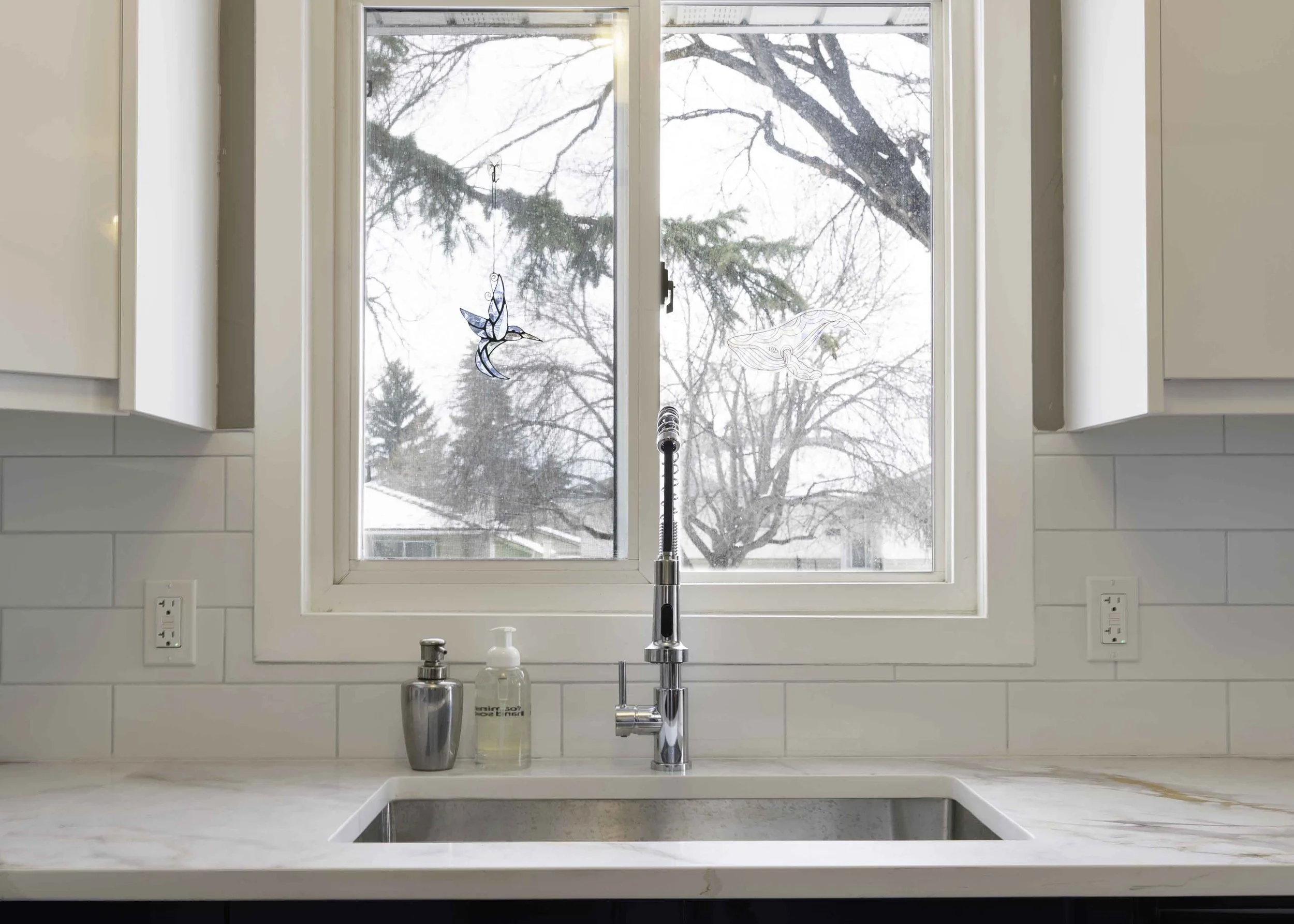 Kitchen sink with a window view of snow-covered trees and rooftops outside, with two decorative glass bird ornaments hanging on the window.