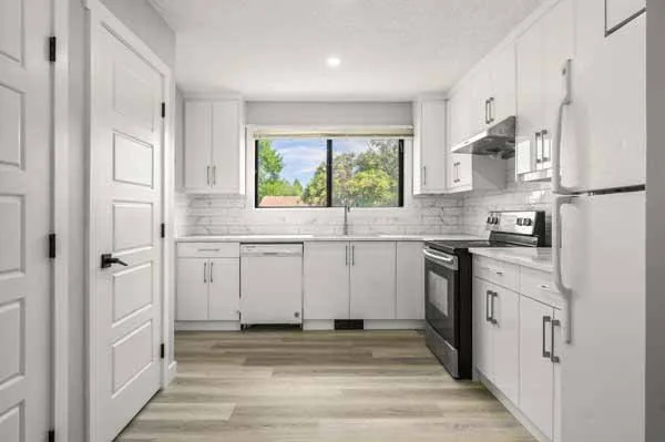 A modern kitchen with white cabinets, a window overlooking green trees, a stove, and white appliances.