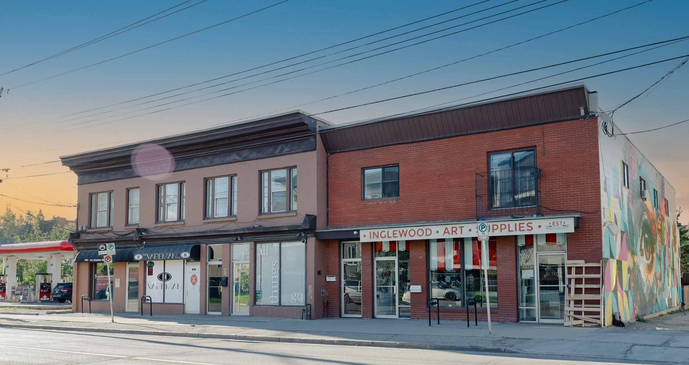 Street view of a two-story commercial building with a mural on the right side, stores on the ground floor, and apartment windows on the upper floor. A gas station is visible on the left, with a sunset sky overhead.