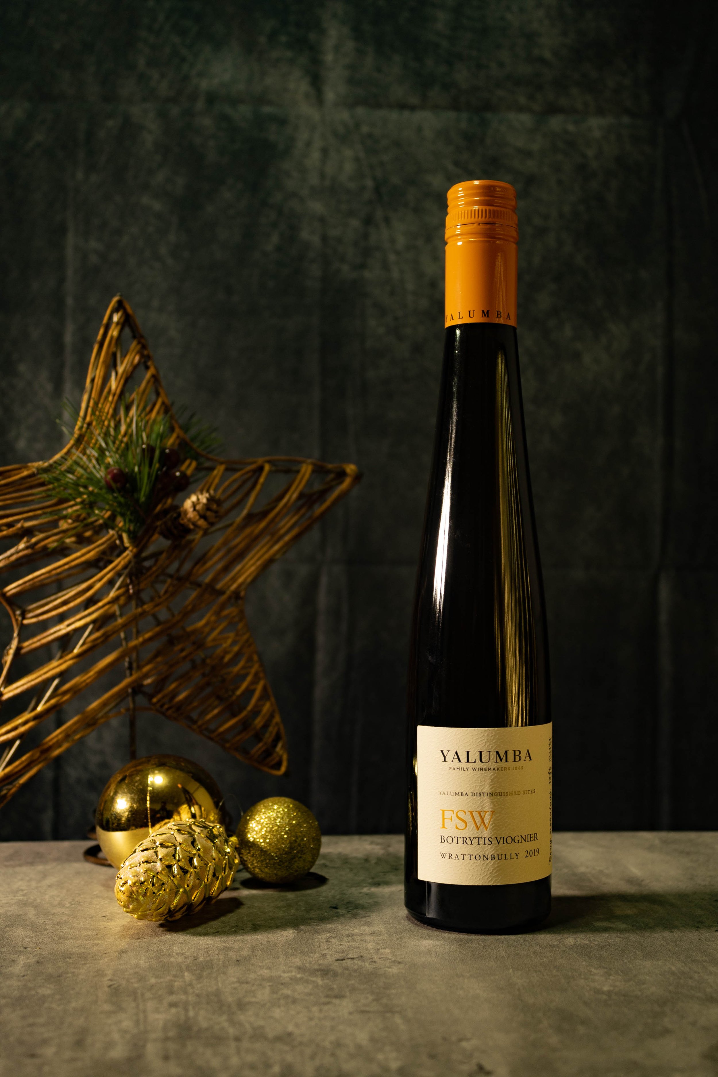 A bottle of Yalumba Botrytis Viognier wine on a gray surface, with a golden star-shaped ornament, two gold Christmas baubles, and a gold pine cone in front, and a woven star decoration on the left.