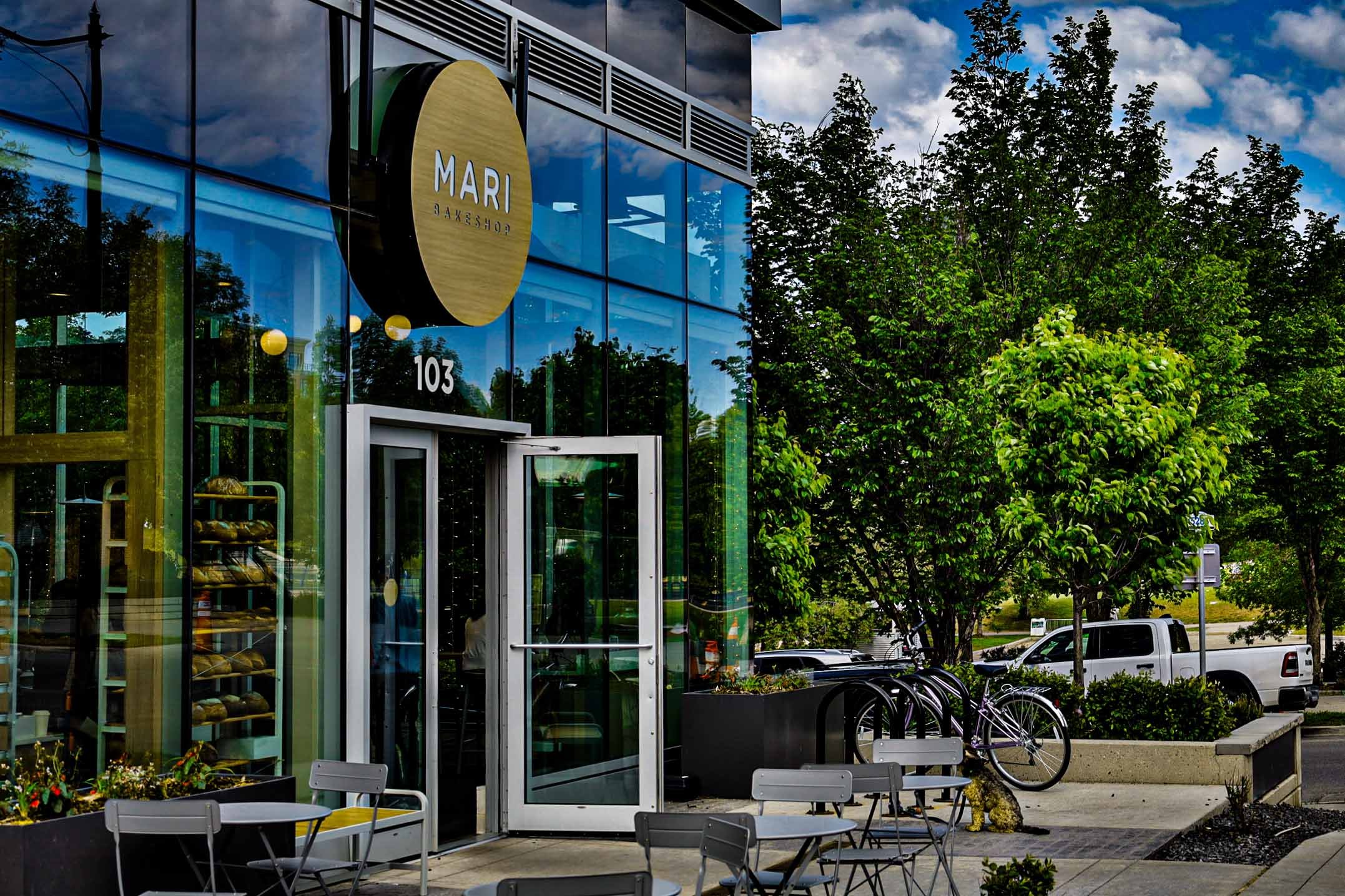 Exterior view of Mari Bakeshop, a modern glass-front bakery, with outdoor seating consisting of metal chairs and tables, bicycles parked nearby, lush green trees, a white pickup truck, and a parking lot under a blue sky with scattered clouds.