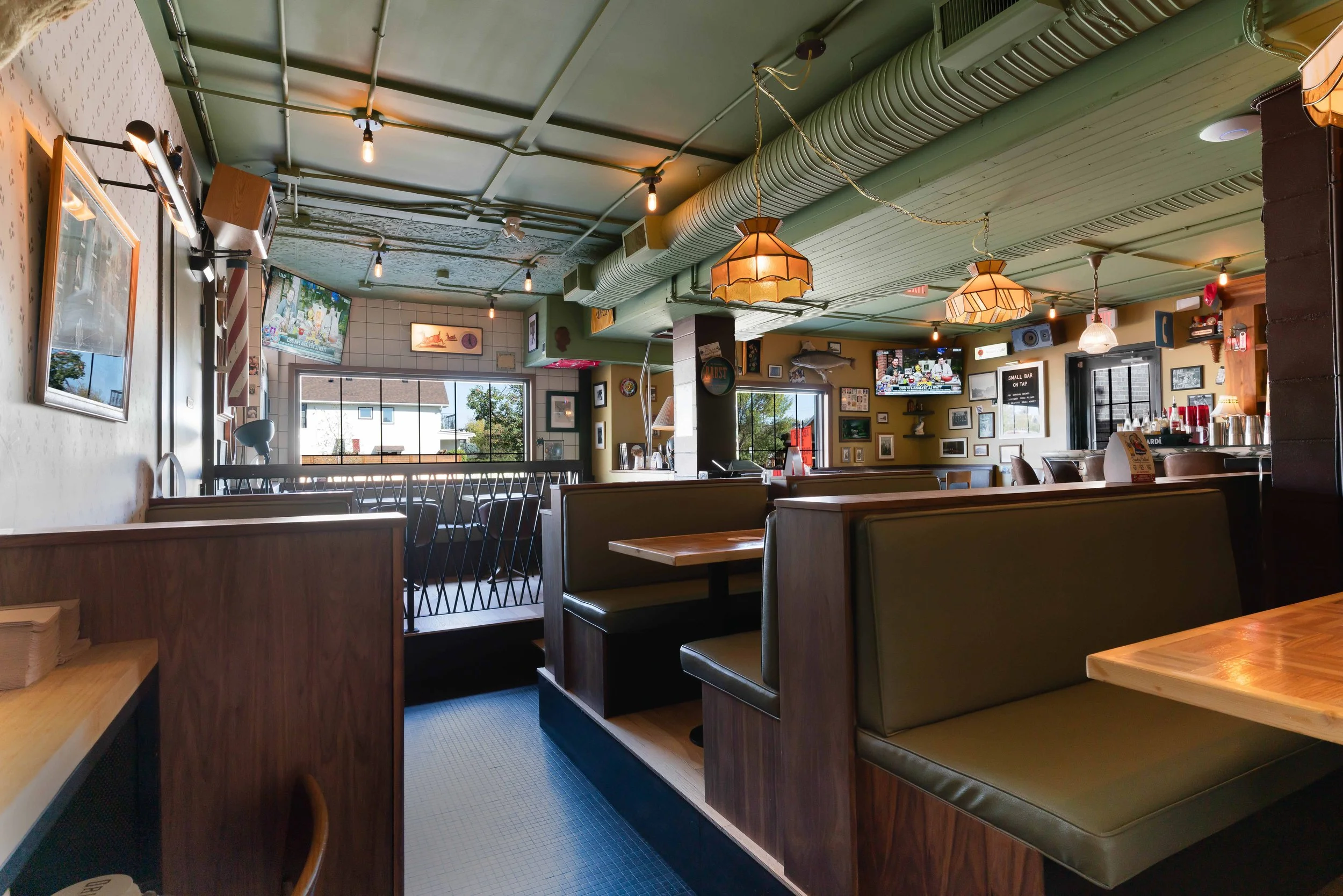 Interior of a cozy restaurant or cafe with green ceiling, wooden booths with green cushions, framed artwork, TV screens, and warm lighting.