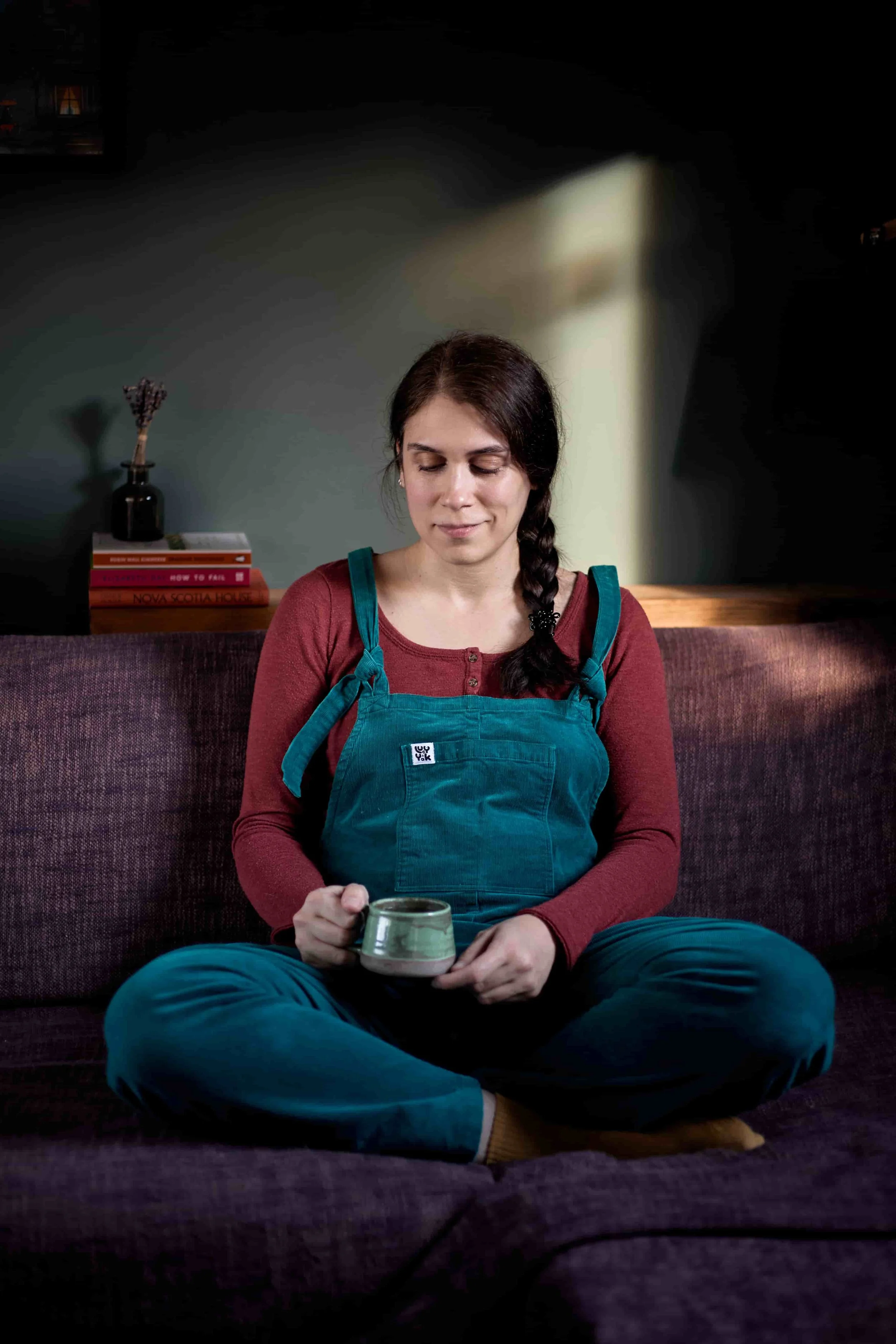 Carina, CBT Therapist, holding a cup of tea, while sitting cross-legged on a sofa in teal corduroy dungarees and a burgundy shirt