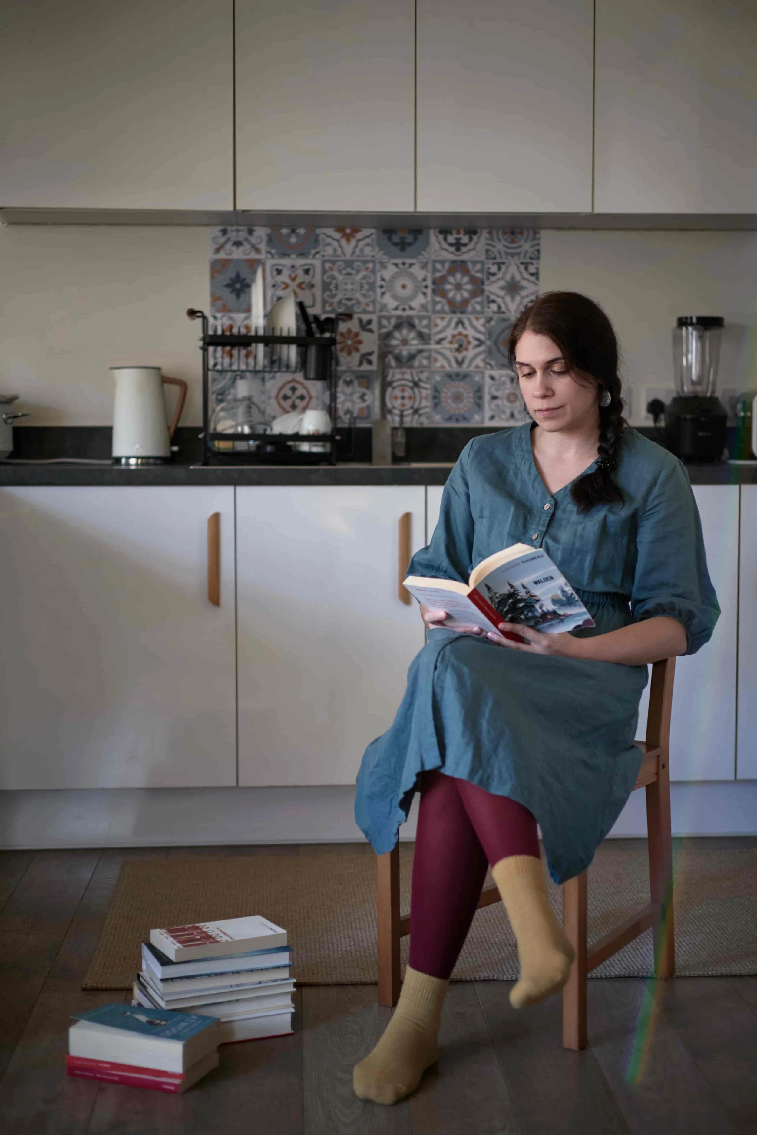 Carina, wearing a teal dress, sitting on a chair cross-leggedin her  kitchen, reading; piles of books on the floor.