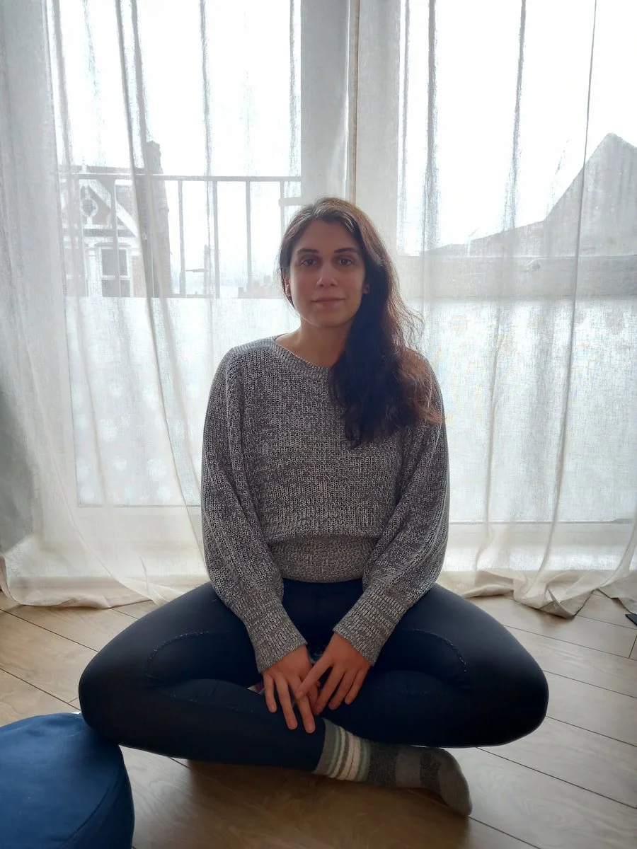 Carina, therapist and mindfulness teacher for high-achievers, sitting cross-legged on a meditation cushion in front of a large window with flowy curtains