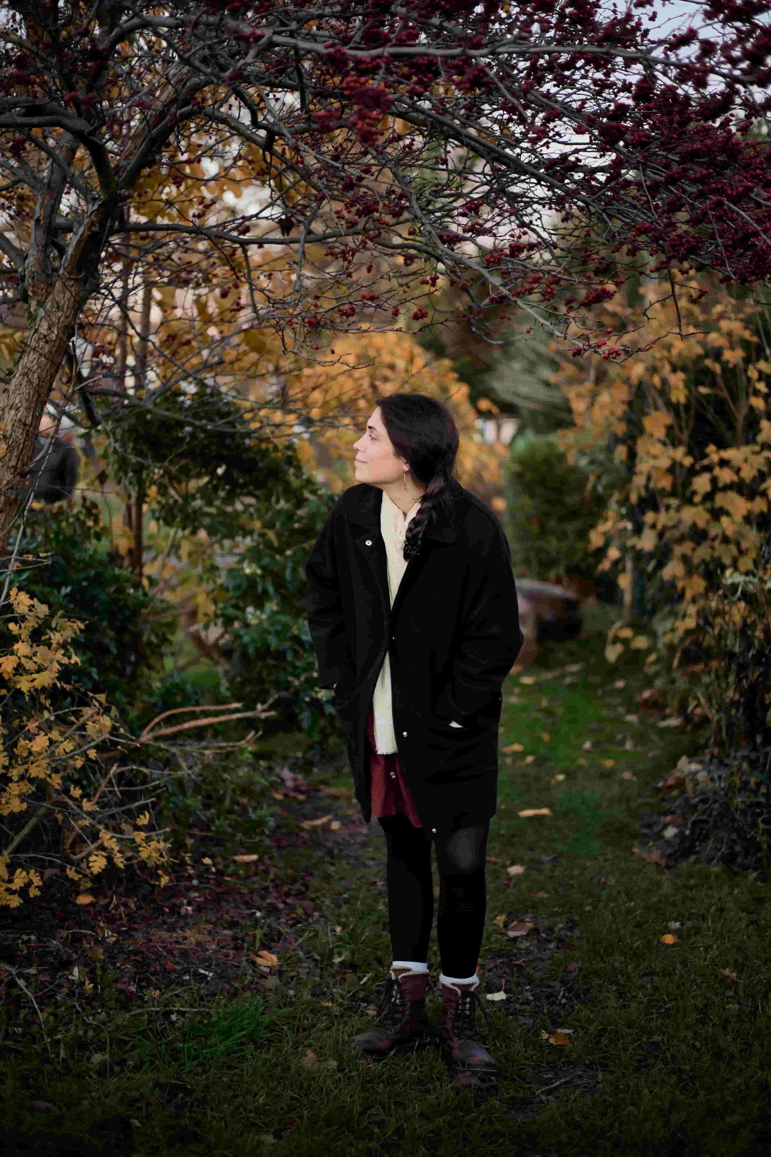 Carina, a dog phobia therapist, standing in a park surrounded by trees and bushes, hands in pockets, looking  to her right, wearing a balck coat, beige scarf, red pinafore dress, balck tights