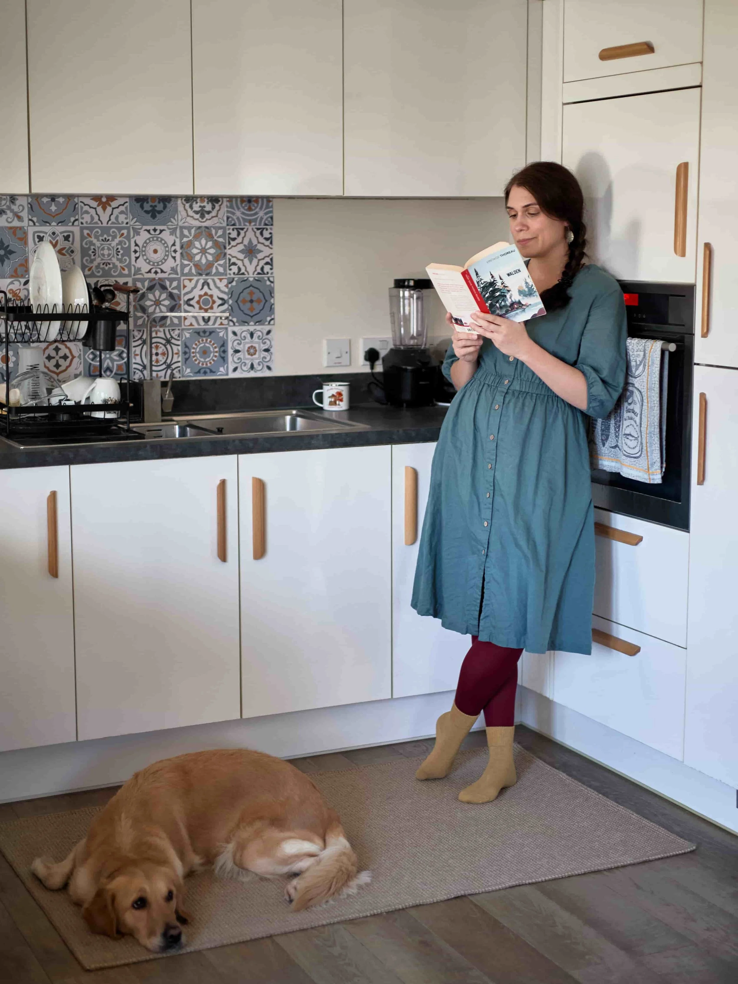 Carina, neuro-affirming therapist, standing in the kitchen holding a book, with her dog, a golden retriever, laying on the floor in front of her