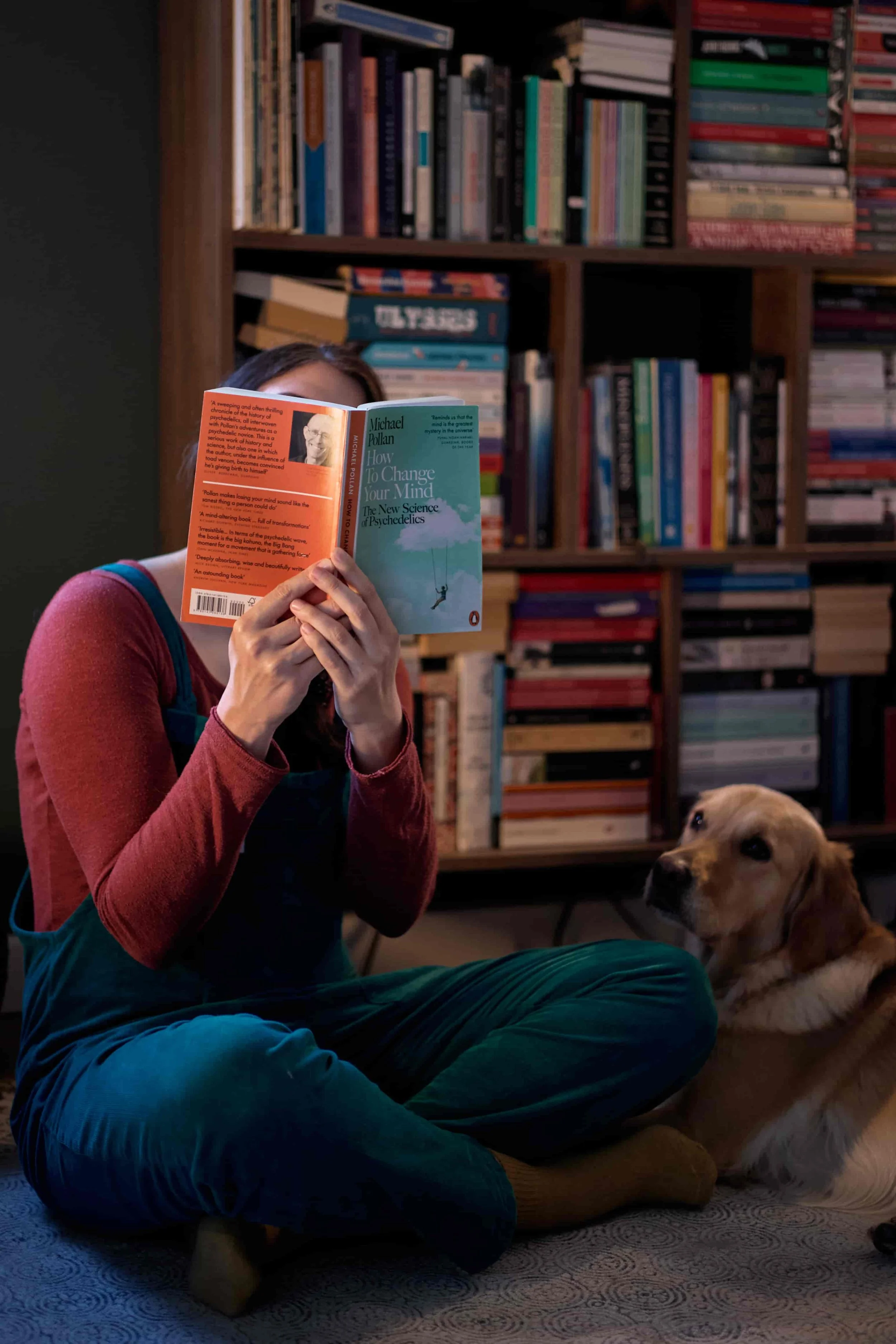 Carina, who offers online CBT therapy in the UK, sitting cross-legged on the floor, holding the book "How to change your mind" in front of her face, her golden retriever dog lying down by her side.
