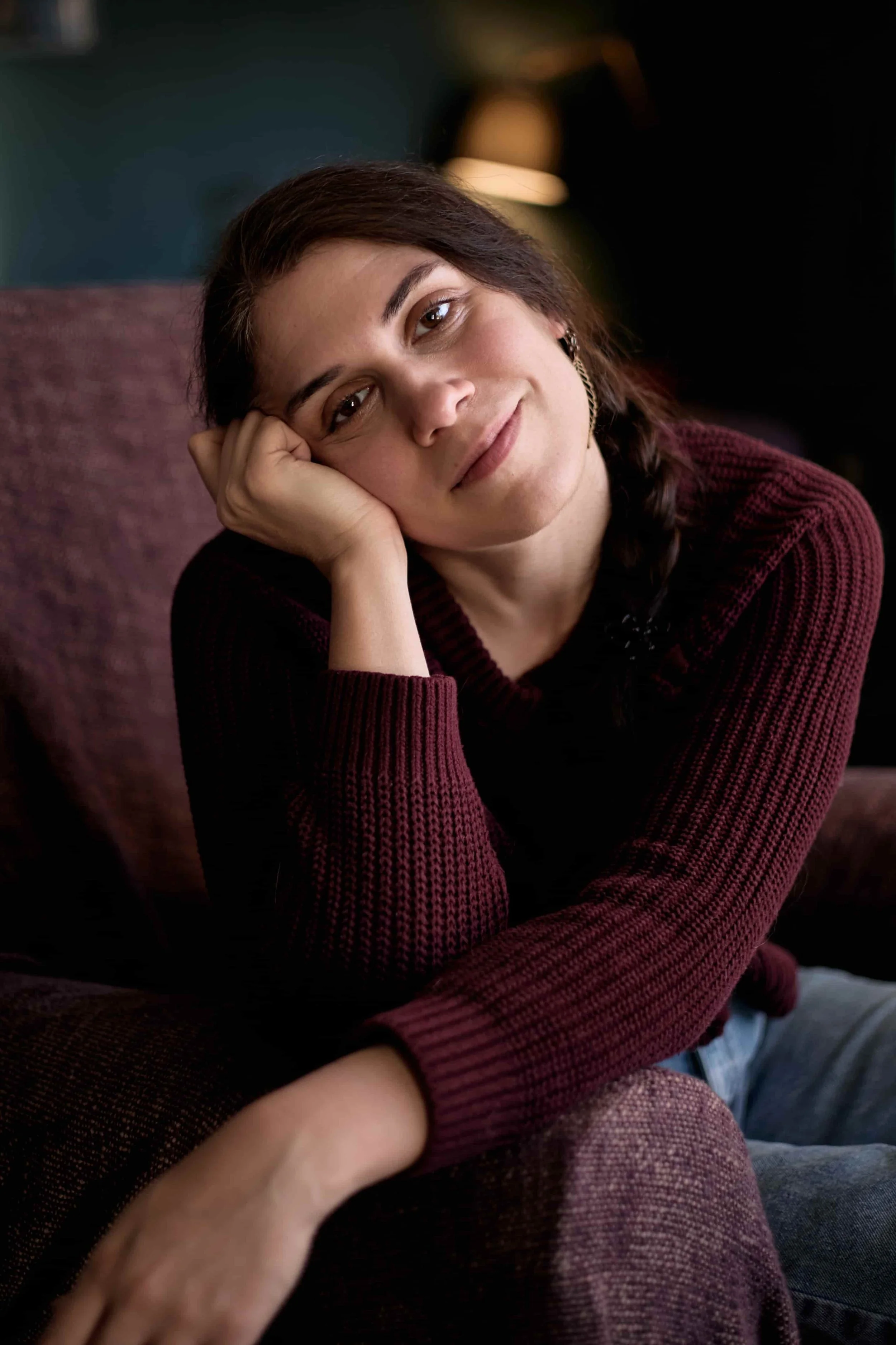 Carina, BABCP-accredited supervisor, wearing a burgundy jumper, sitting on armchair, resting head in hand, smiling.