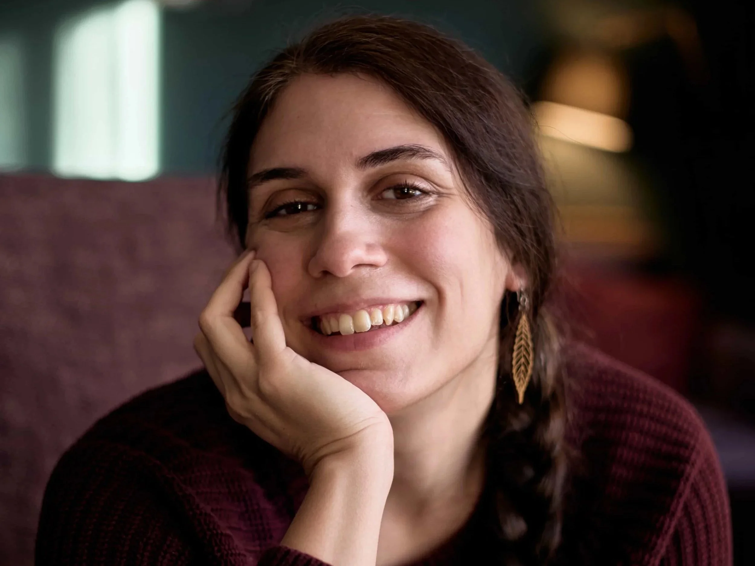 Carina, a therapist who works with self-doubting overachievers, wearing a burgundy jumper, leaf-shaped dangly earrings, and her hair in a side-braid, resting her head in her hand and smiling warmly at the camera