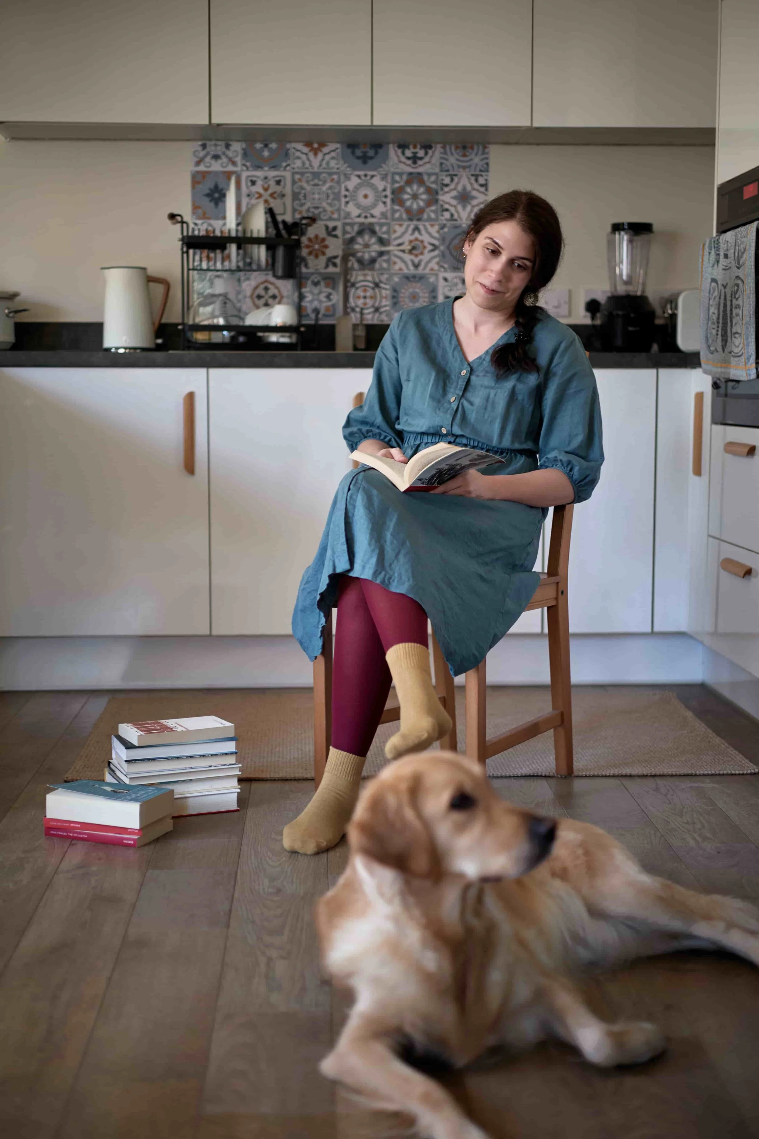 Carina, neuro-affirming therapist, sitting on a chair in the kitchen holding a book, looking at her dog, a golden retriever