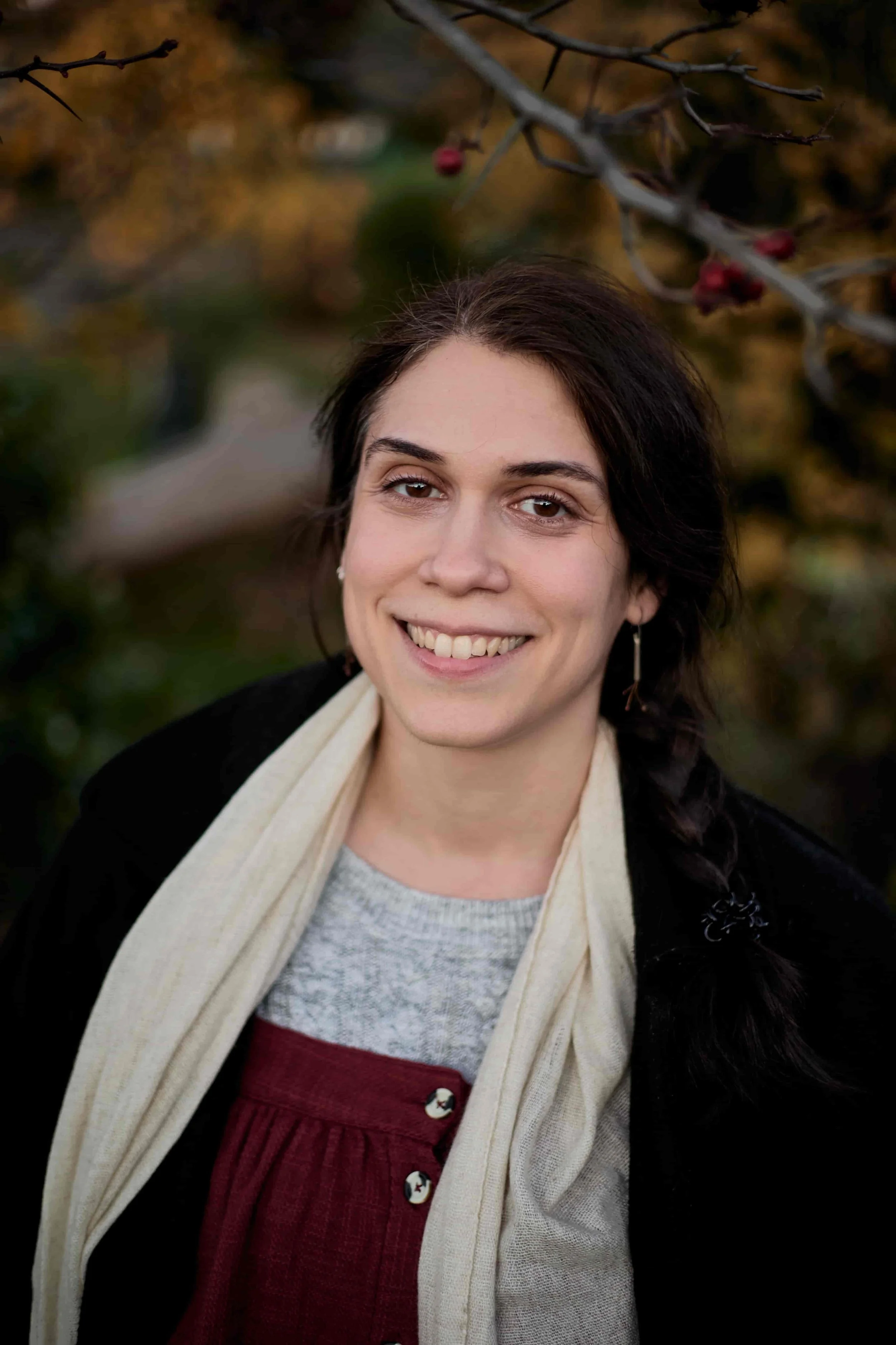 Carina, LGBTQIA+-affirming therapist, smiling at the camera in a park, wearing a pinafore dress and jumper, beige scarf and black coat