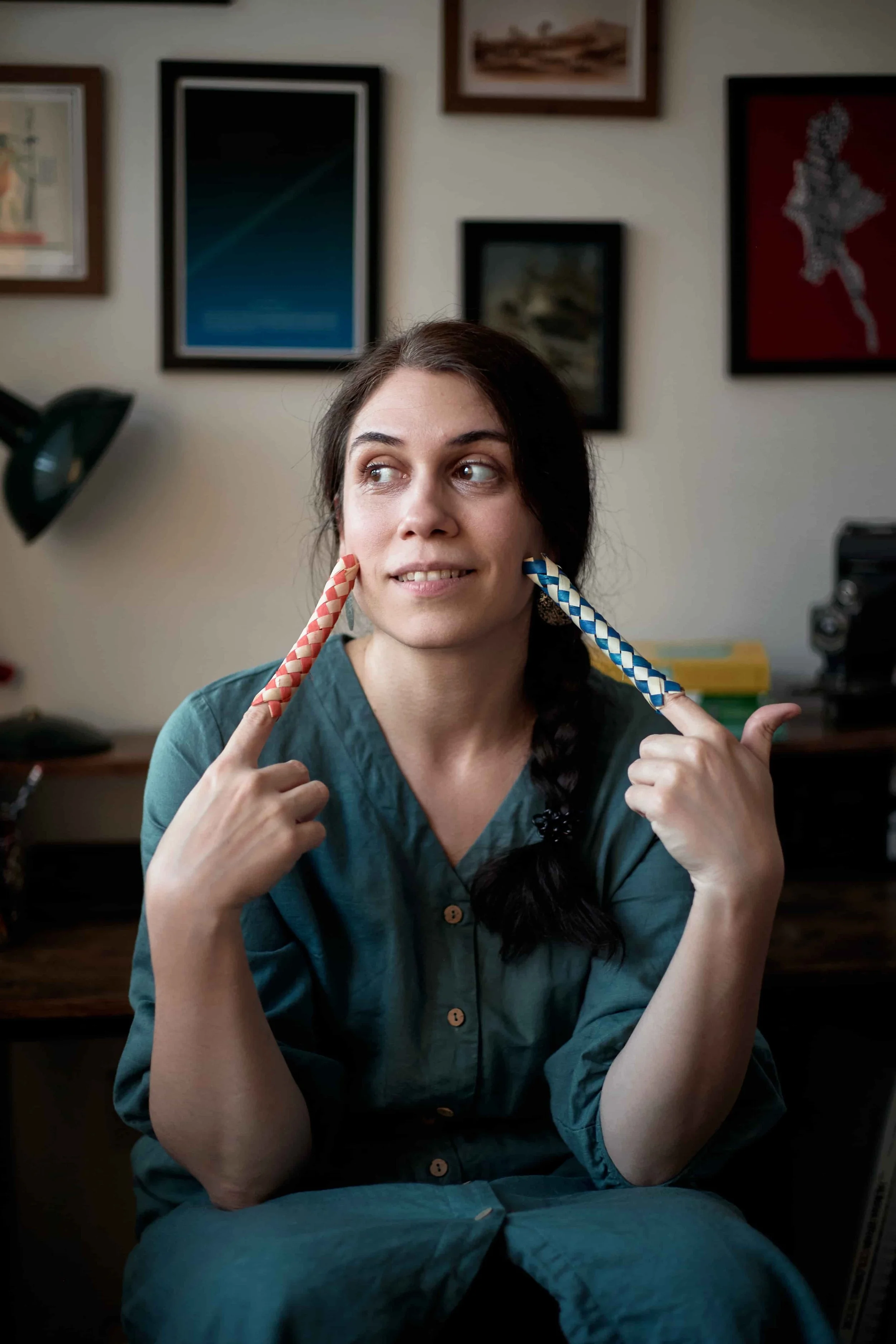 Carina wearing a teal dress sitting in front of a desk with framed prints in the background, wwith Chinese fingertraps on each index finger, touching her face with them