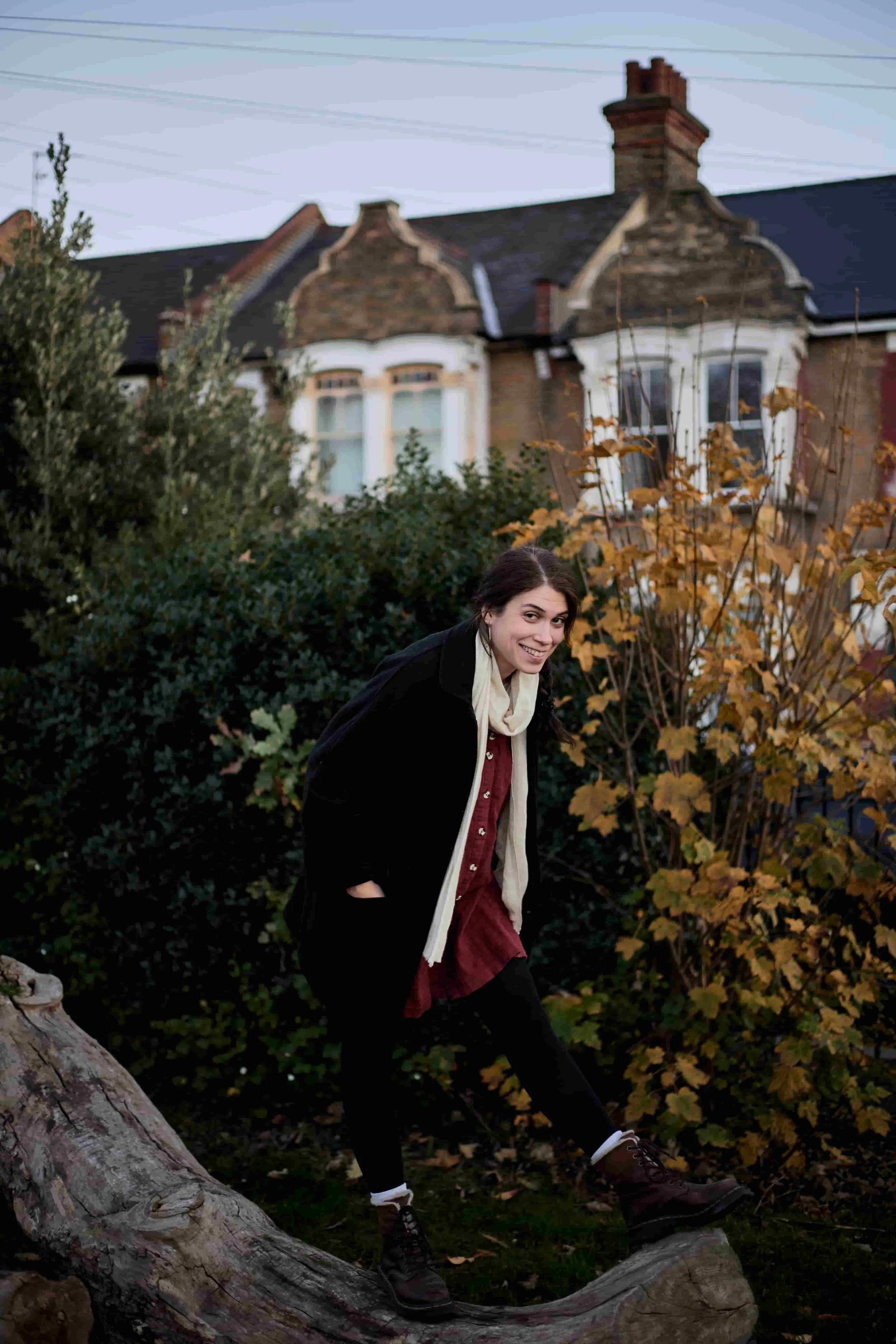 Carina, a neuroa-ffirming therapist, wearing a burgundy pinafore dress, beige scarf, black coat, and black tights, standing in a funny posture on a fallen tree in a park.