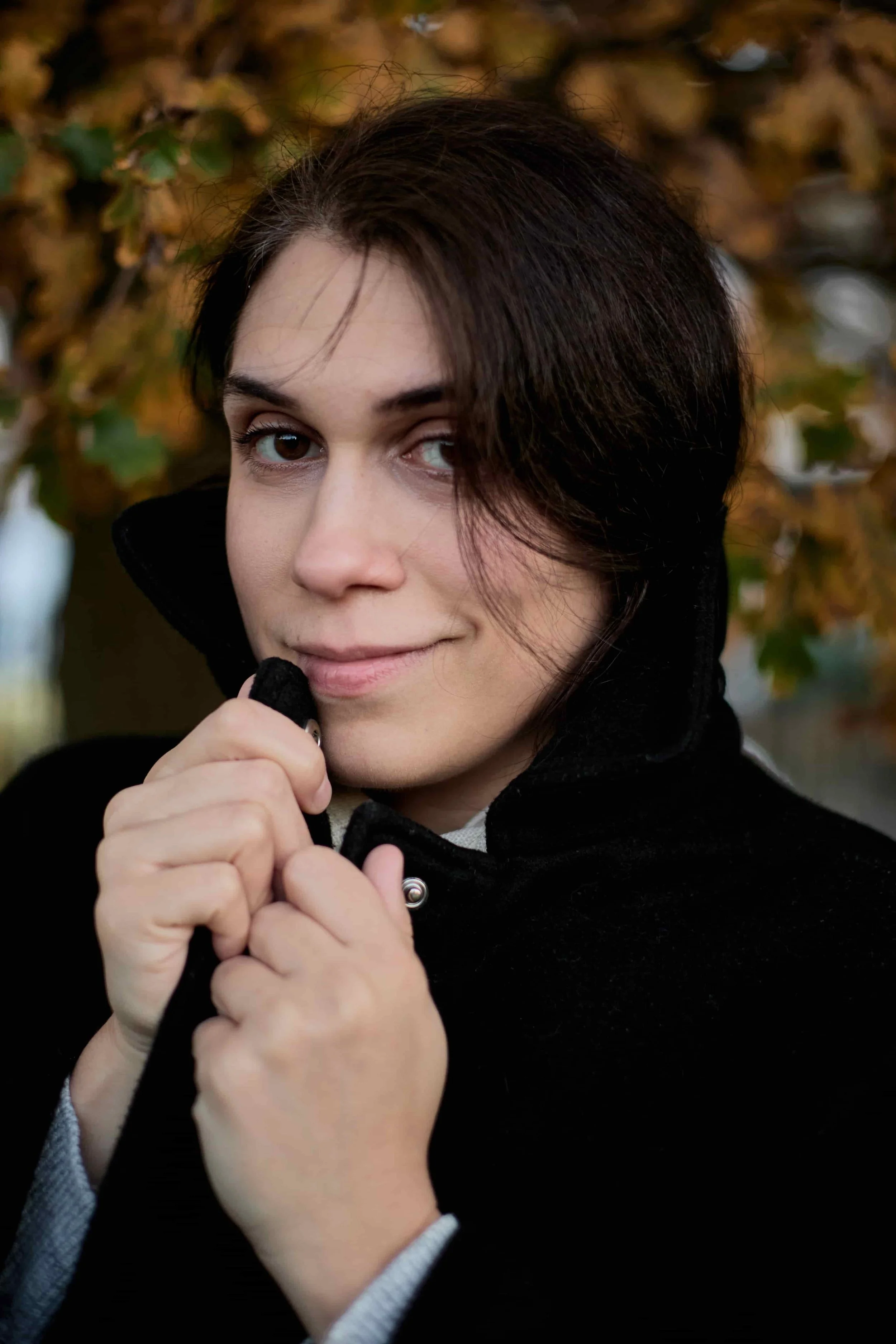 Carina, a CBT Therapist for Overthinkers, clasping her black coat outside, background of autumn leaves - a headshot