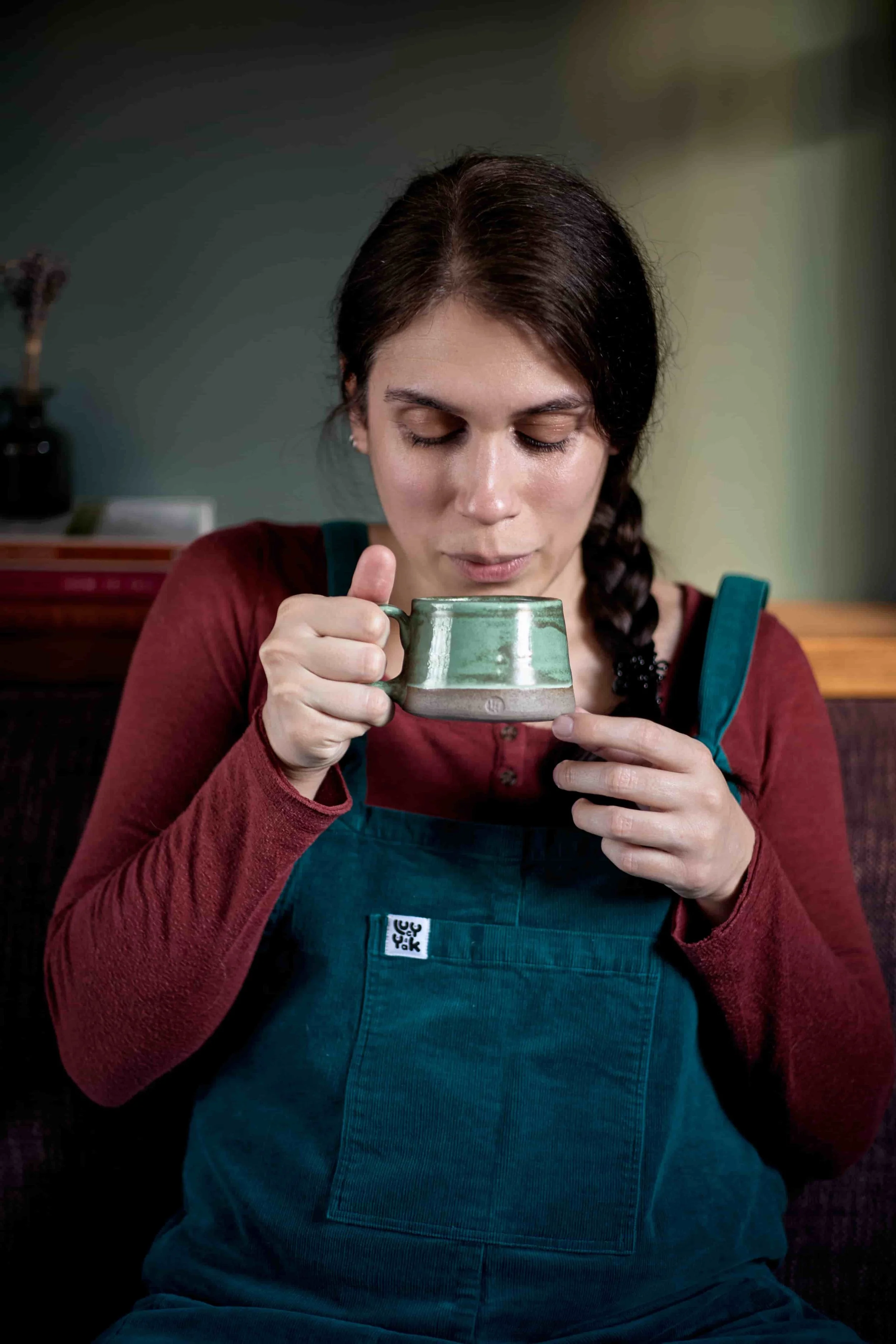 Carina, CBT Therapist, blowing on a hot cup of tea, while sitting on a sofa in teal corduroy dungarees and a burgundy shirt