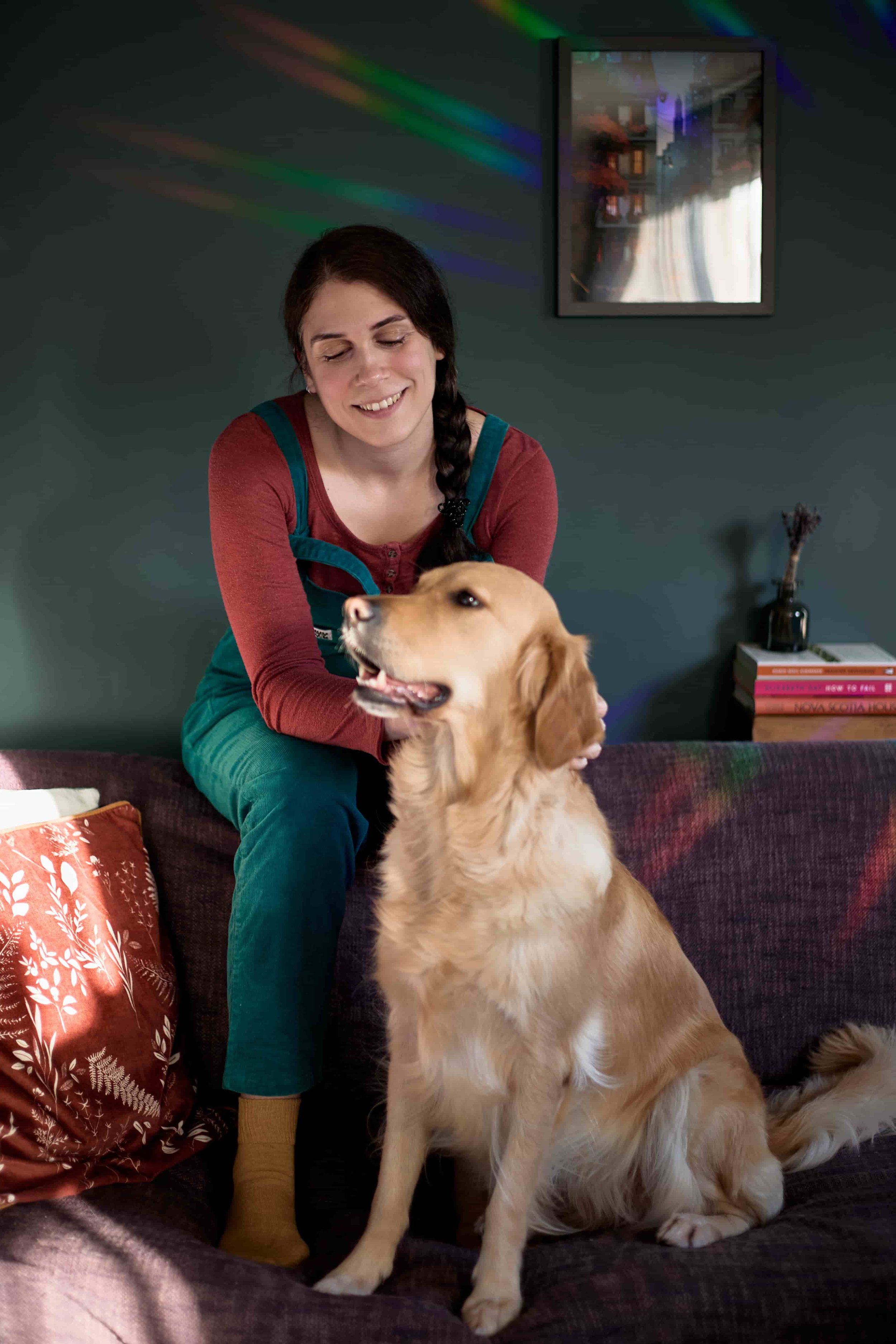 Carina, a BABCP-accredited supervisor, sitting on the backrest of a sofa wearing teal dungarees and a burgundy shirt, petting her dog, a golden retriever