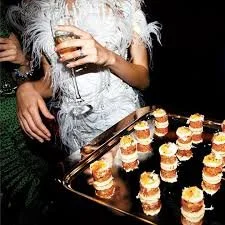 A person holding a glass of champagne next to a tray of small, layered appetizers or desserts, likely at a party or celebration.