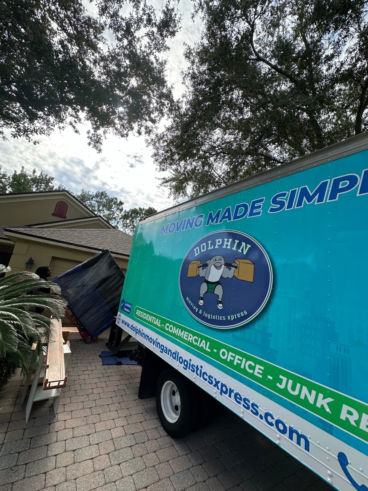 A moving company truck with the logo 'Dolphin Moving & Logistics Xpress' and slogan 'Moving Made Simple,' parked on a brick driveway next to a house with trees and clouds overhead.
