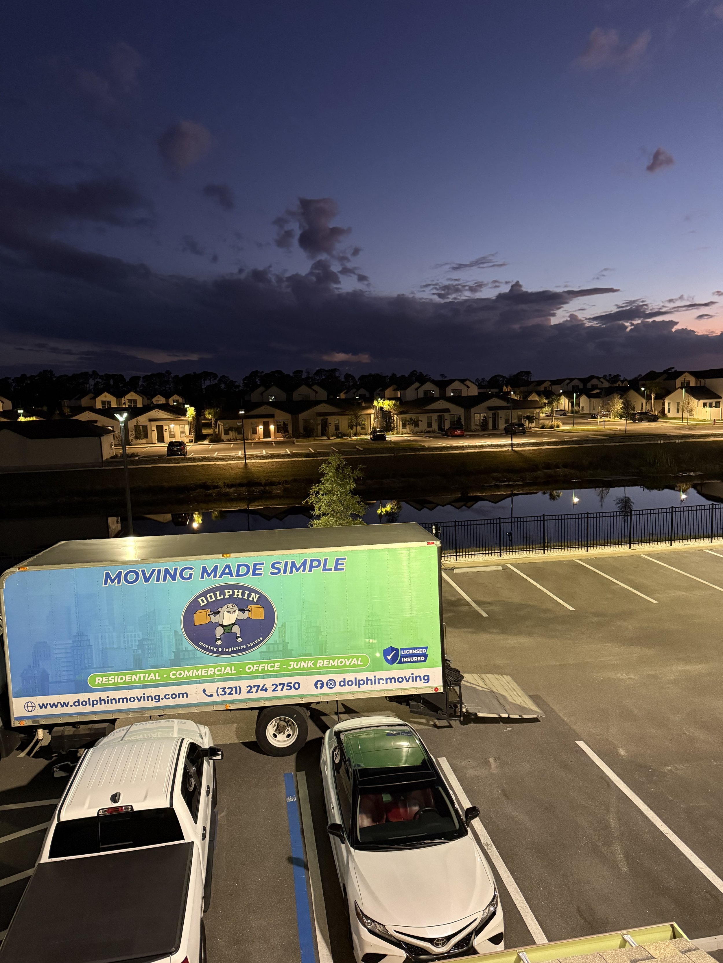 A large moving truck labeled 'Dolphin Moving' parked in an parking lot at dusk. The truck has a blue and green design and the company's website and contact info on the side. Two cars are parked beside the truck, and a small tree is visible behind it. In the background, there are residential houses with illuminated windows and a body of water reflecting the lights and sky.