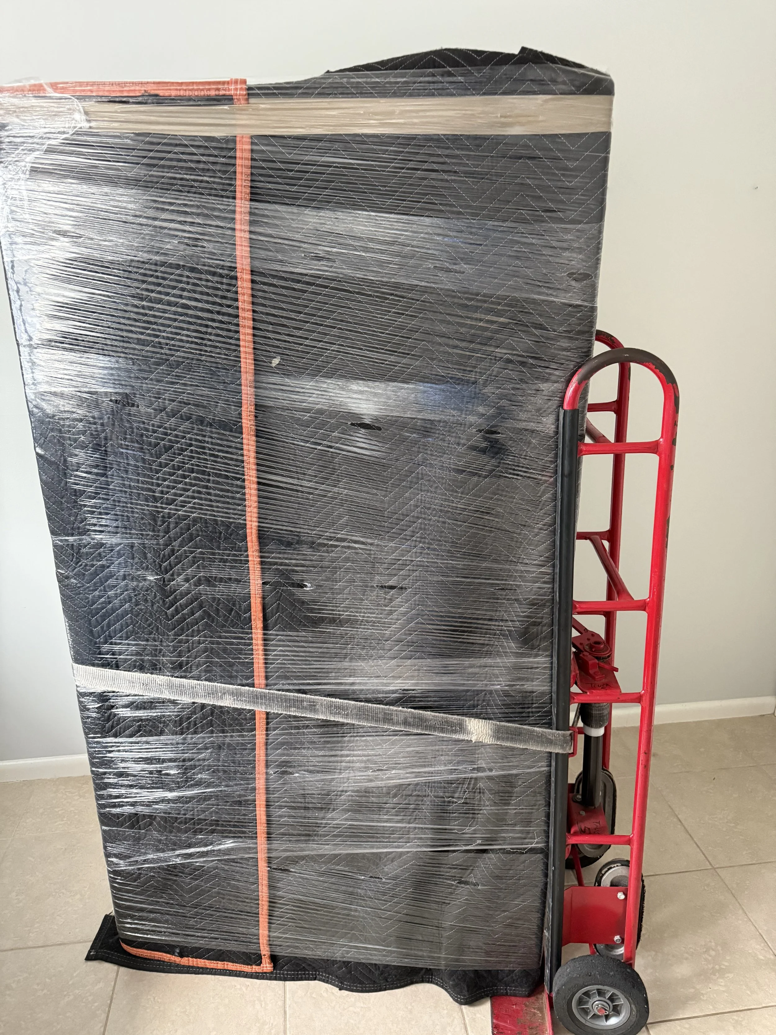 A large wrapped item on a red hand truck, covered with transparent plastic wrap, positioned indoors against a plain white wall.