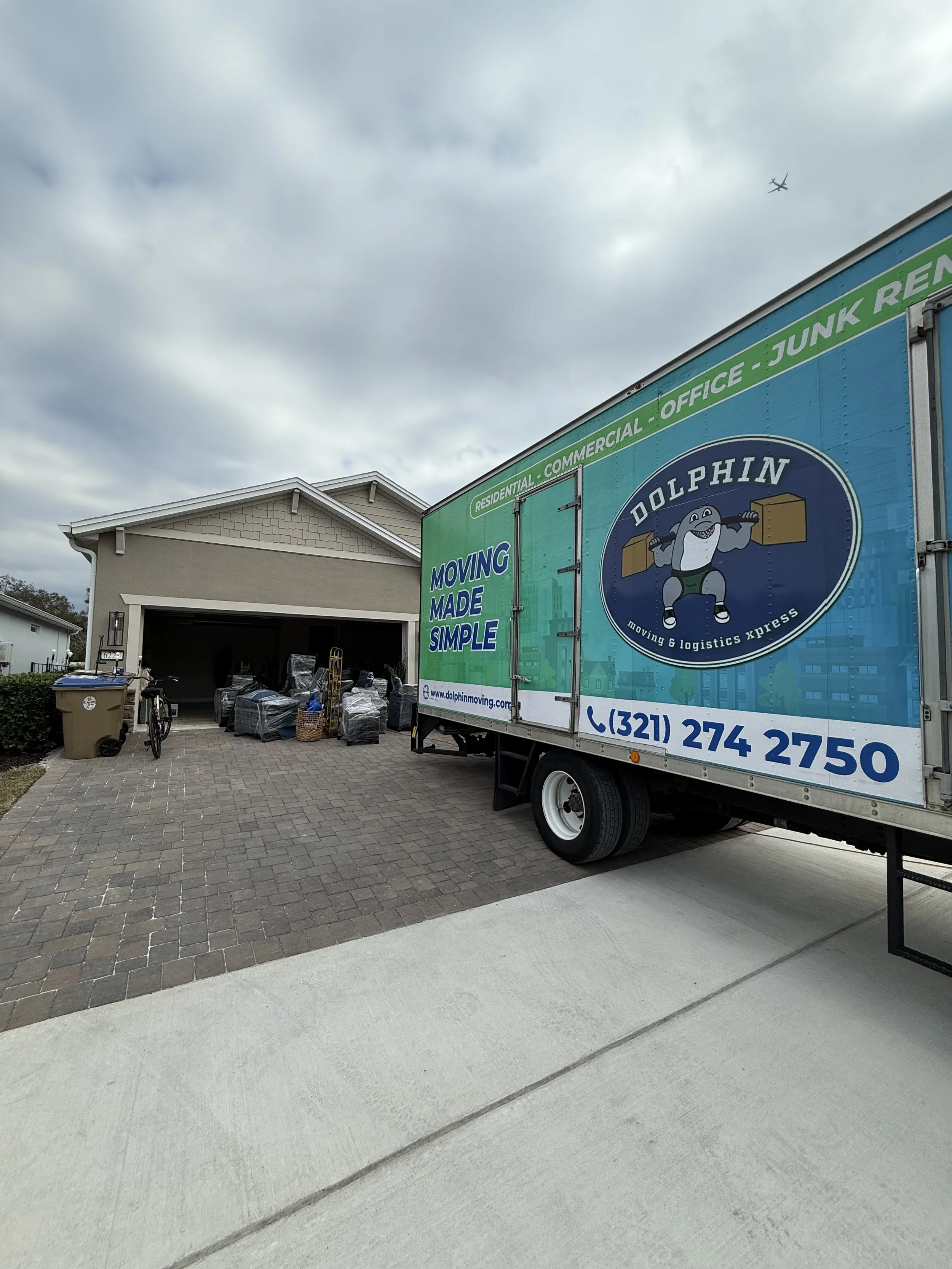 Moving truck parked in driveway with furniture and boxes near garage of a house. Overcast sky with airplane visible in the distance.