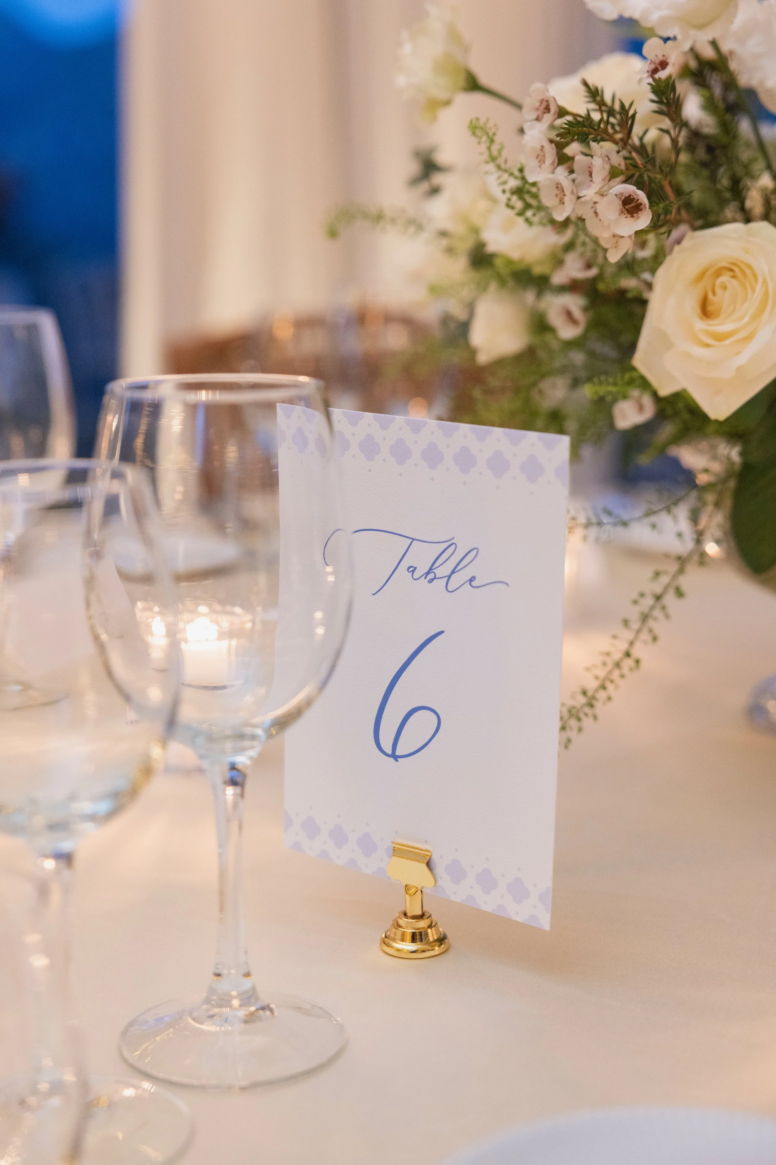 A table number card with the number 6, surrounded by several empty wine glasses, with a floral centerpiece including white roses and greenery in the background.