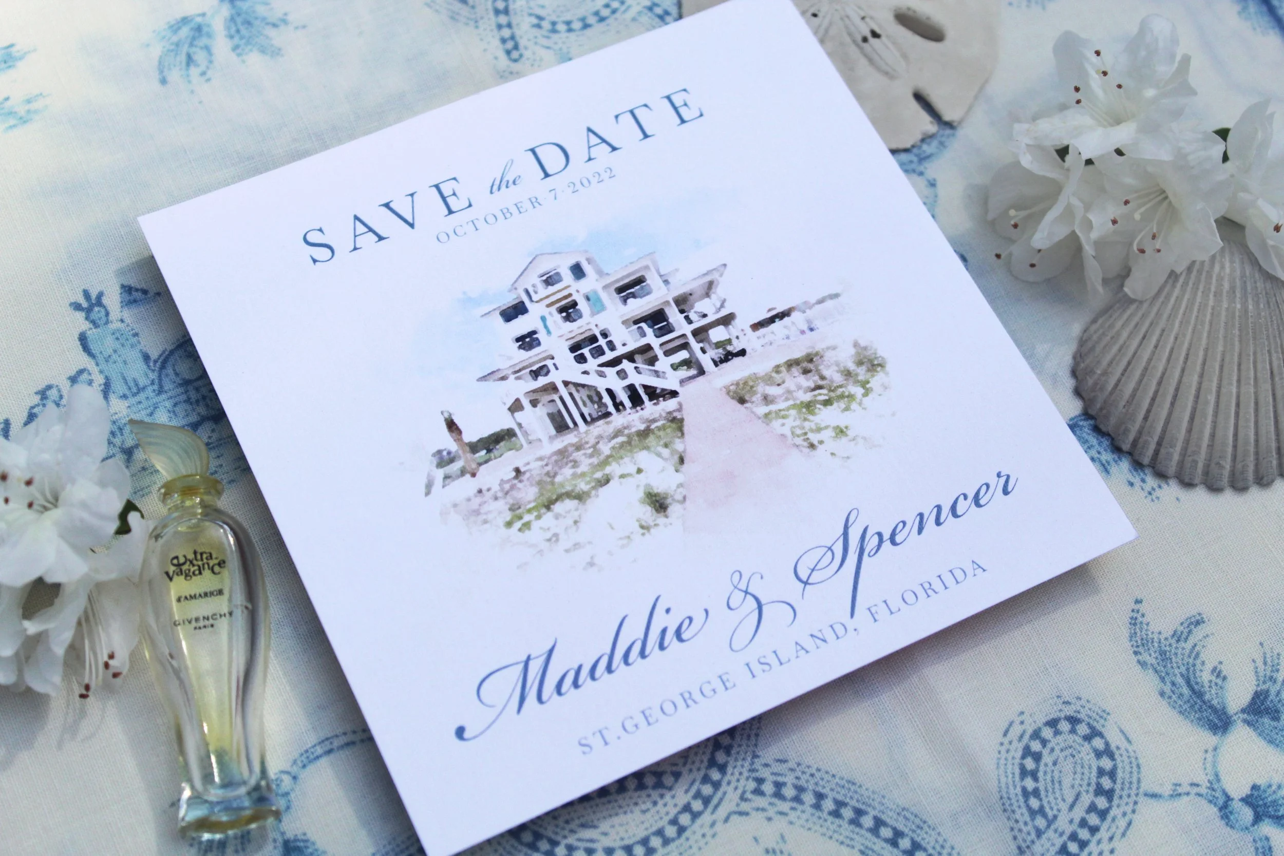 Save the date wedding announcement card for Maddie and Spencer, October 7, 2022, featuring an illustration of a beach house and located on a table with white flowers, a seashell, and a small bottle of perfume.