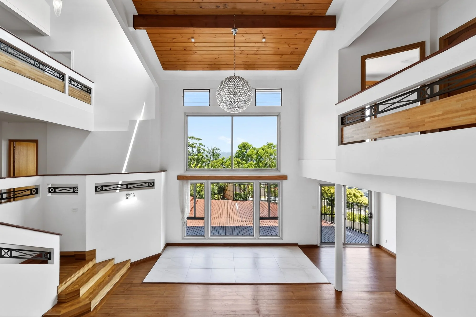 Interior view of a modern, spacious living room with large windows, wooden floors, white walls, and a high ceiling with a wooden panel and chandelier.