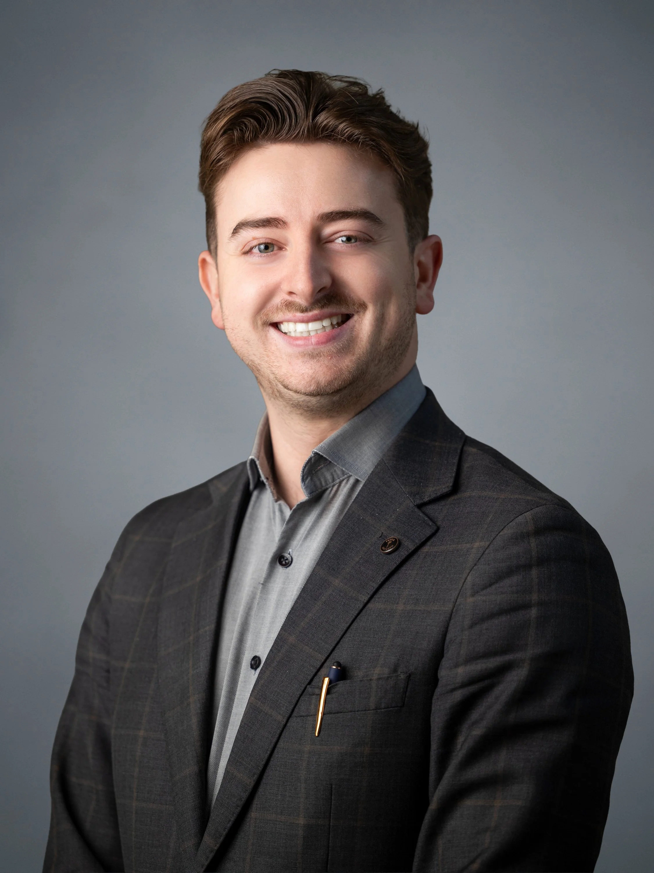 A portrait of a young man with brown hair, blue eyes, and light skin, smiling and wearing a dark plaid blazer over a gray dress shirt, against a gray background.