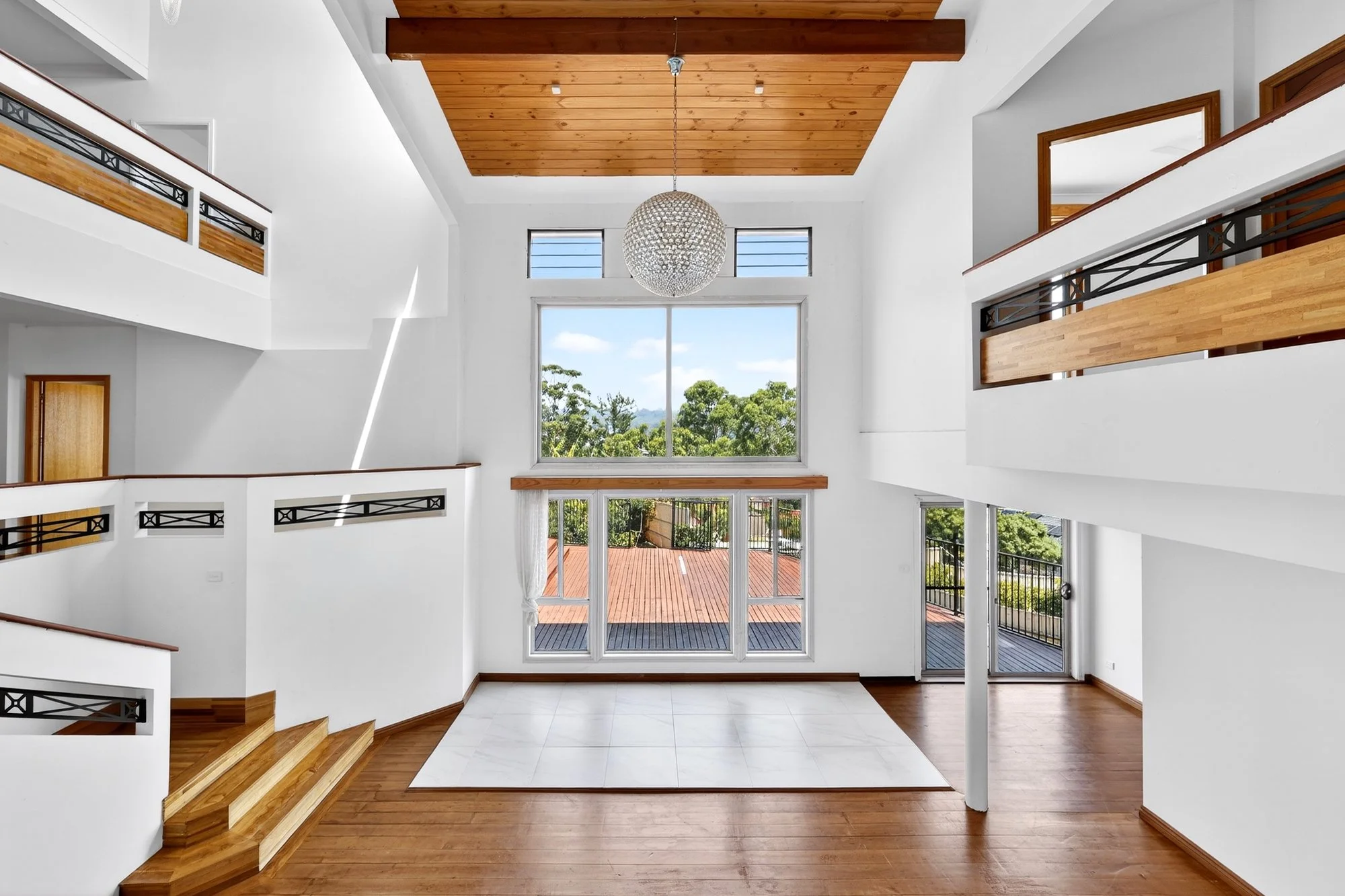Interior of a modern, spacious house with white walls, large windows, wooden accents, and a high ceiling with a chandelier.