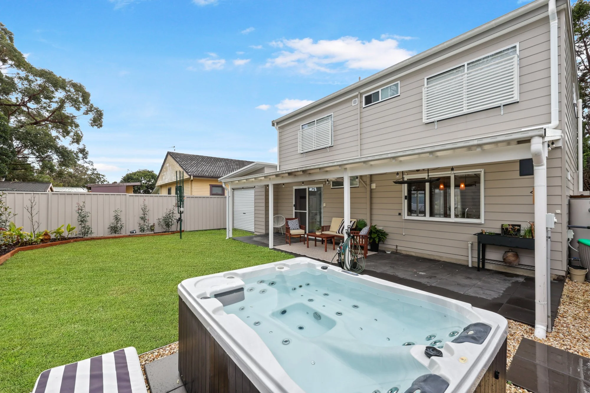 Backyard with a hot tub, green lawn, patio area, house with beige siding, white fences, and a small garden bed with plants. Blue sky with some clouds.