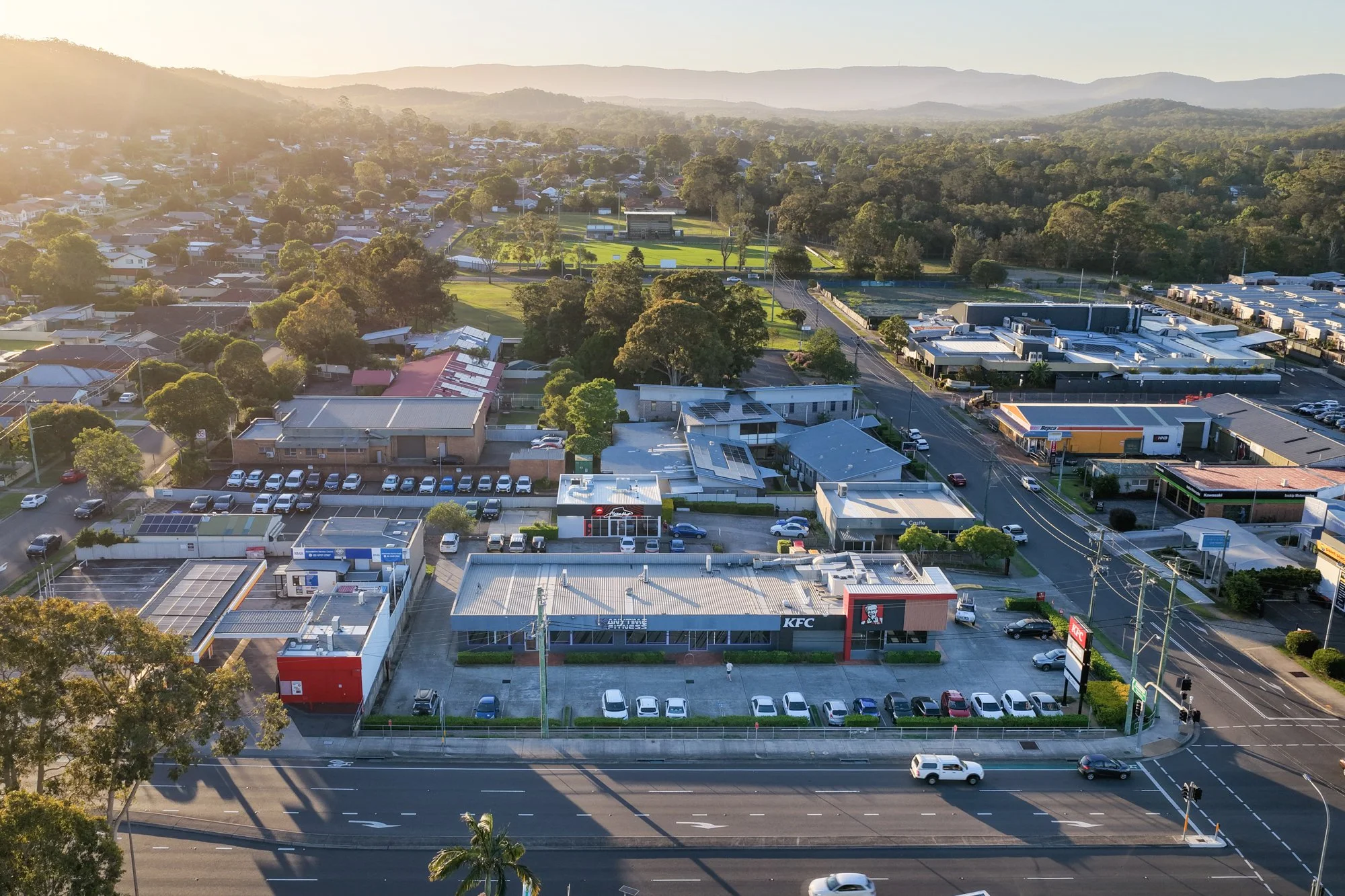 An aerial view of a shopping center with a KFC restaurant, parking lots, and surrounding commercial and residential buildings, with a green park area and hilly landscape in the background during sunset.