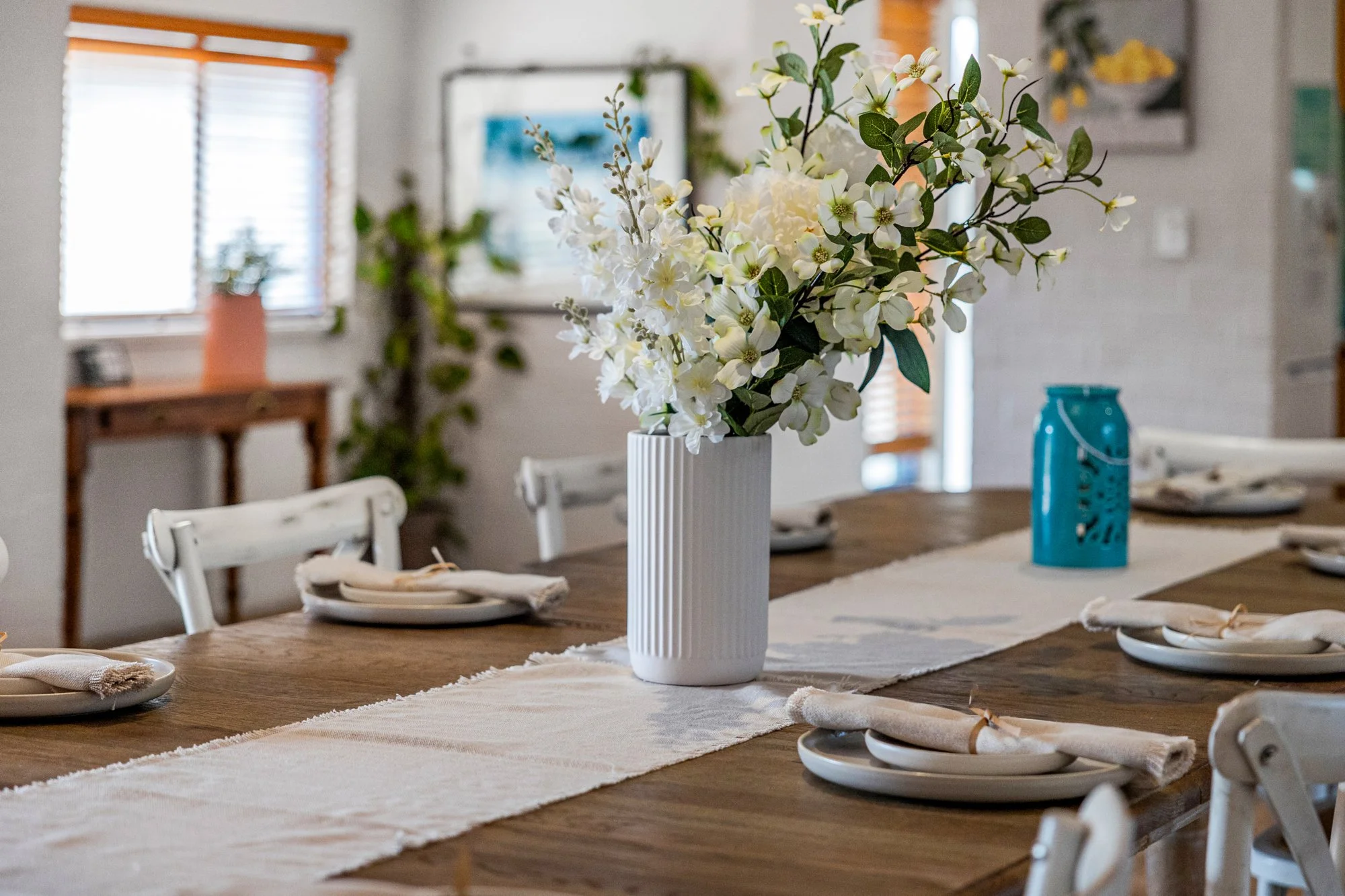A dining table decorated with a white vase holding white flowers, a table runner, and neatly folded napkins on white plates in a bright room with windows and green plants.