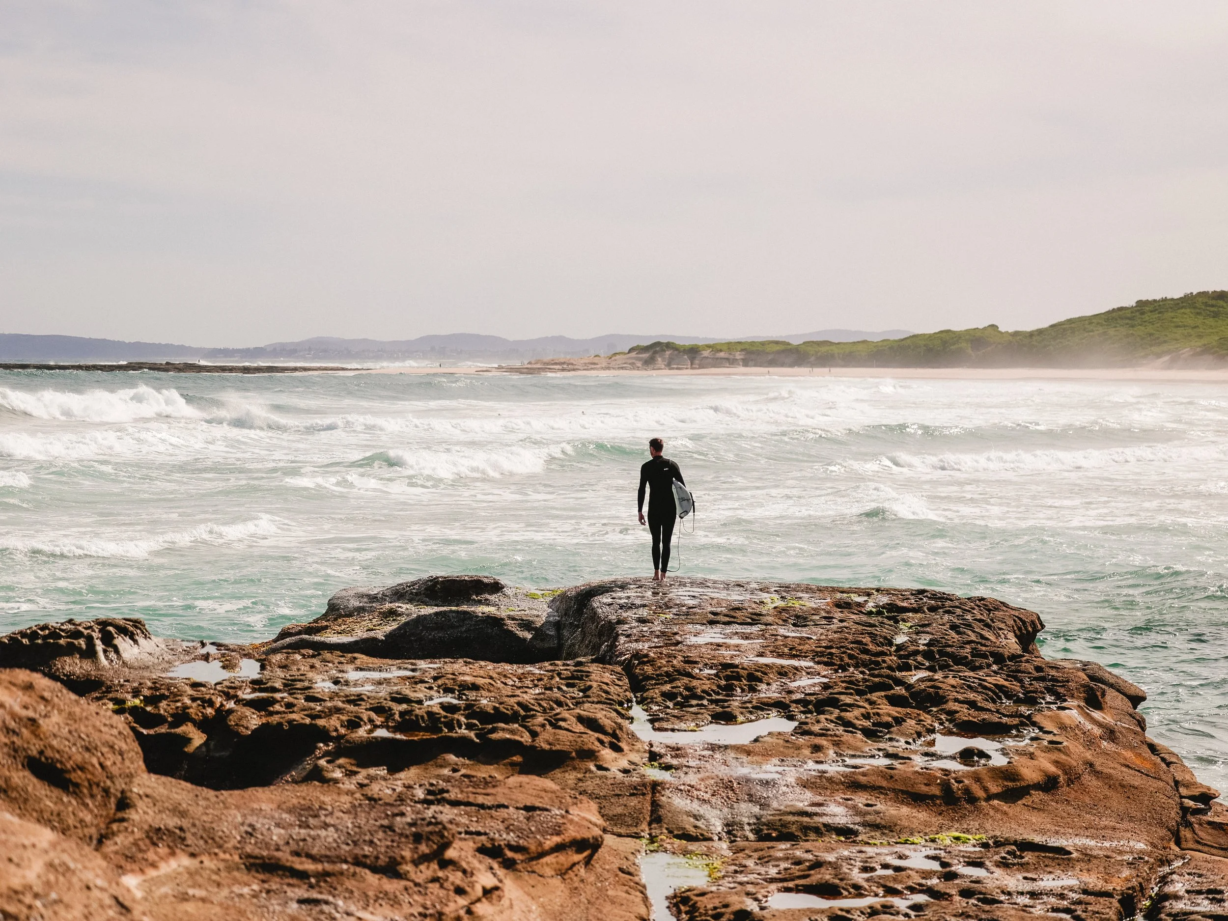A person in a wetsuit holding a surfboard walking on rocks towards the ocean waves at a beach.