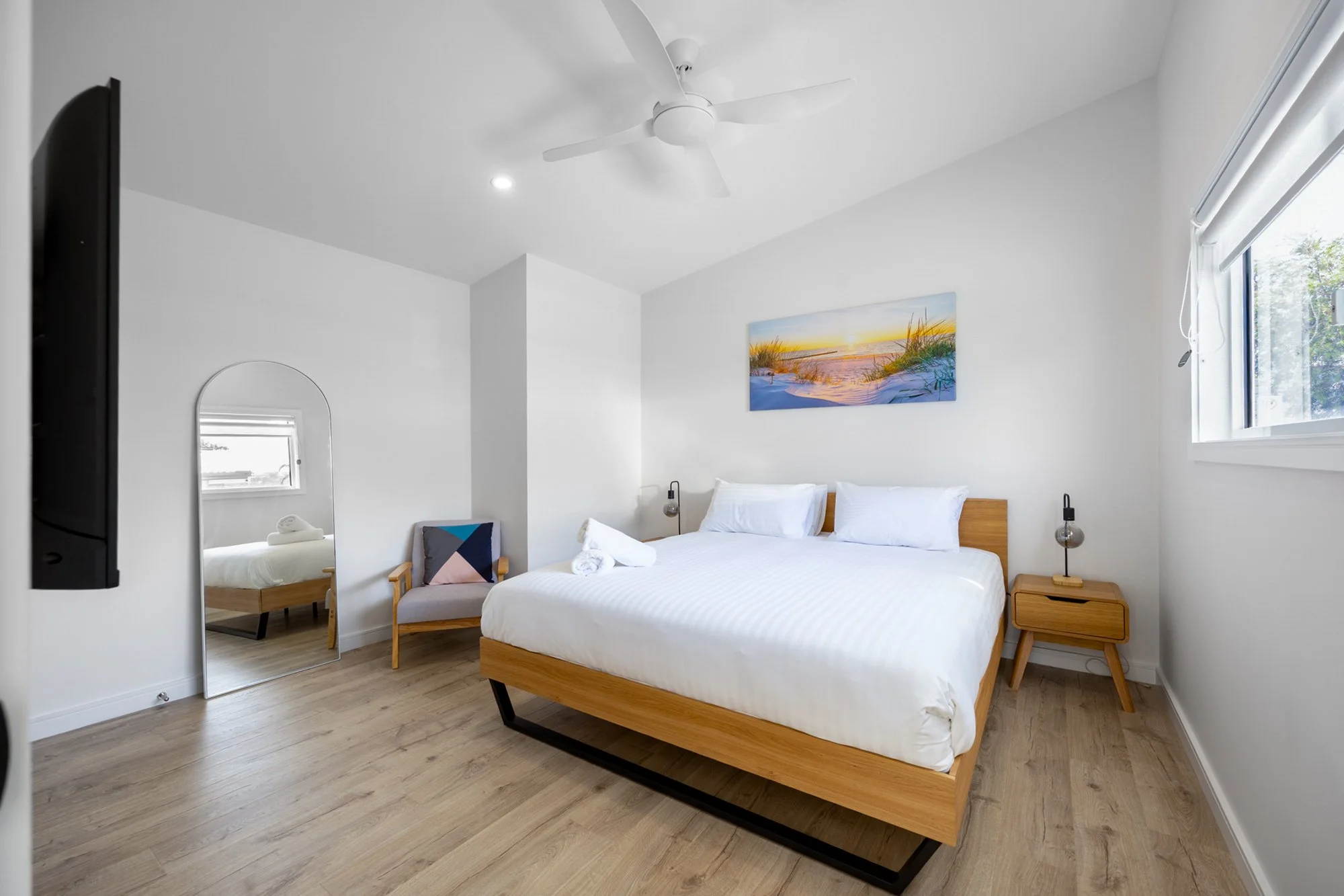 Modern bedroom with wooden bed frame, white bedding, minimalist nightstands, wall art, and a ceiling fan. Features a large mirror and armchair beside a window with blinds.