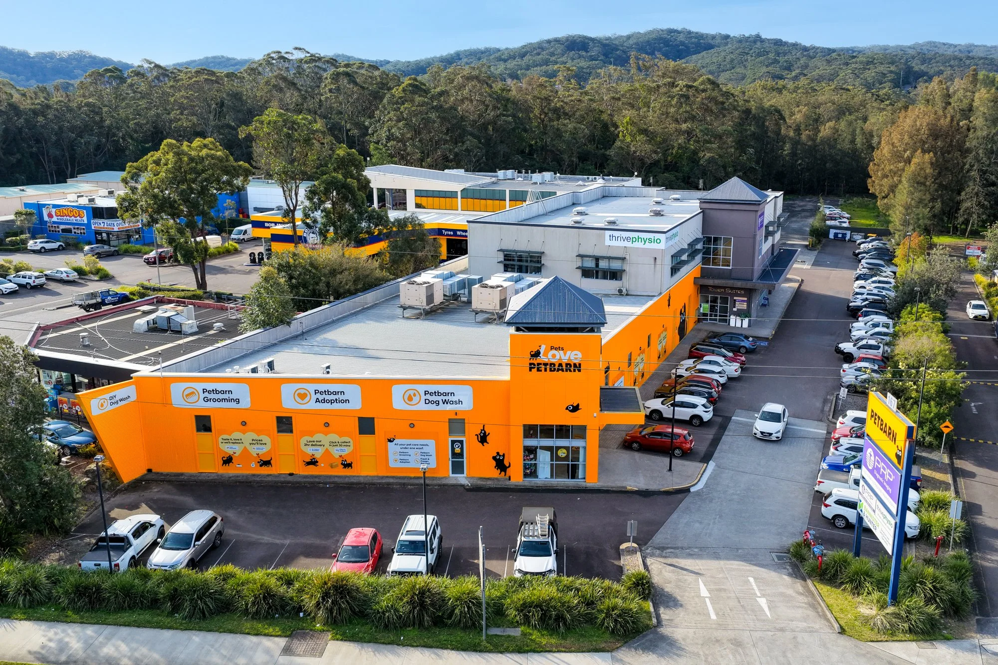 Aerial view of a commercial building housing a pet grooming, adoption, and dog wash business with orange exterior, surrounded by parking lots filled with cars, and a nearby shopping center with trees and a hilly landscape in the background.