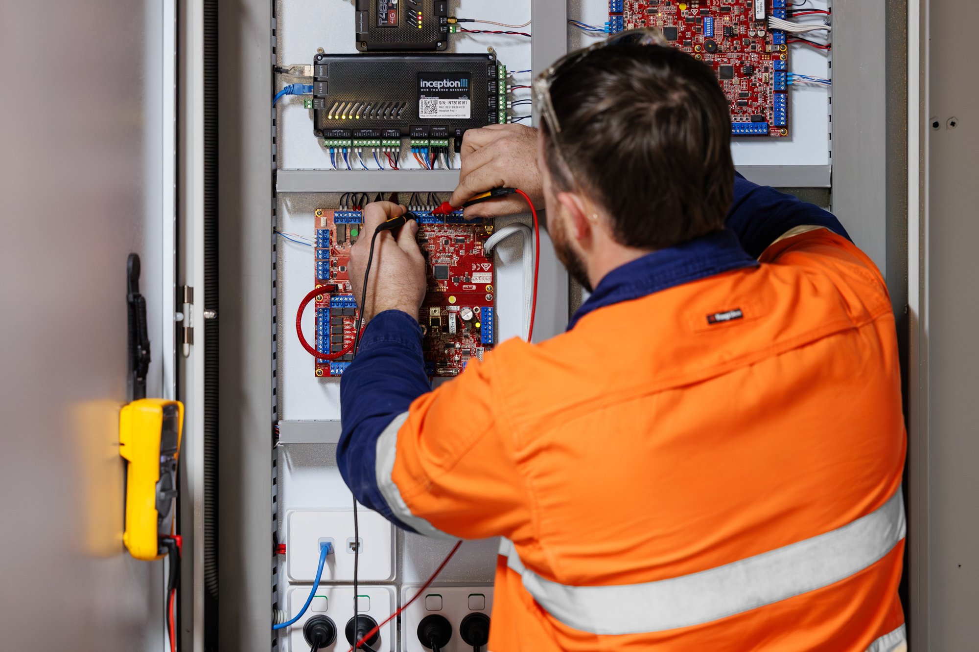 A worker in an orange safety vest and glasses is repairing or maintaining an electrical control panel with various circuit boards and wiring inside a metal enclosure.