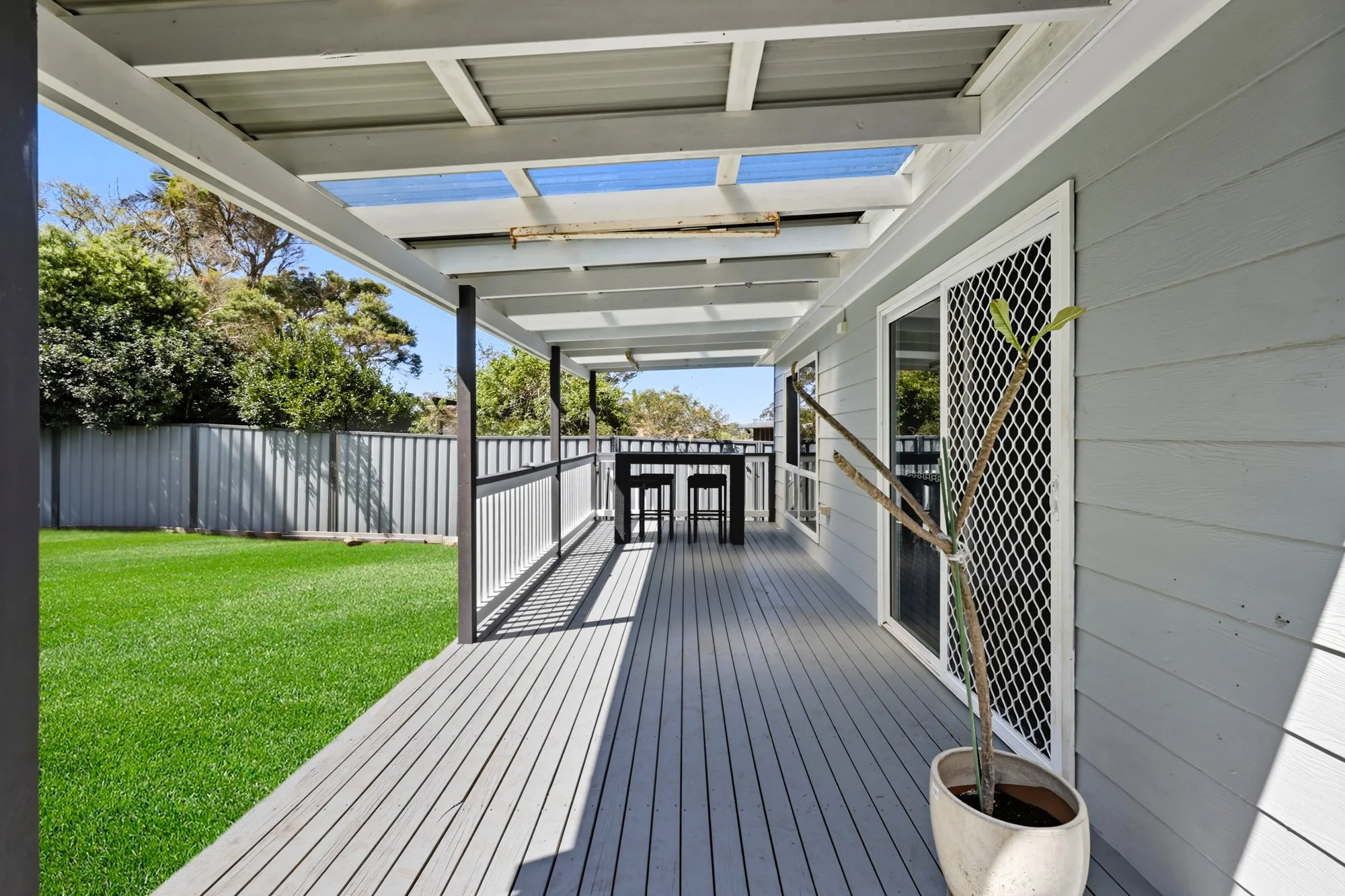 An outdoor balcony with wooden flooring and a white house exterior. There is a potted plant with a thick stem and sparse leaves. The balcony has a black railing, two bar stools and a small table at the far end. A fence and trees are visible in the background.