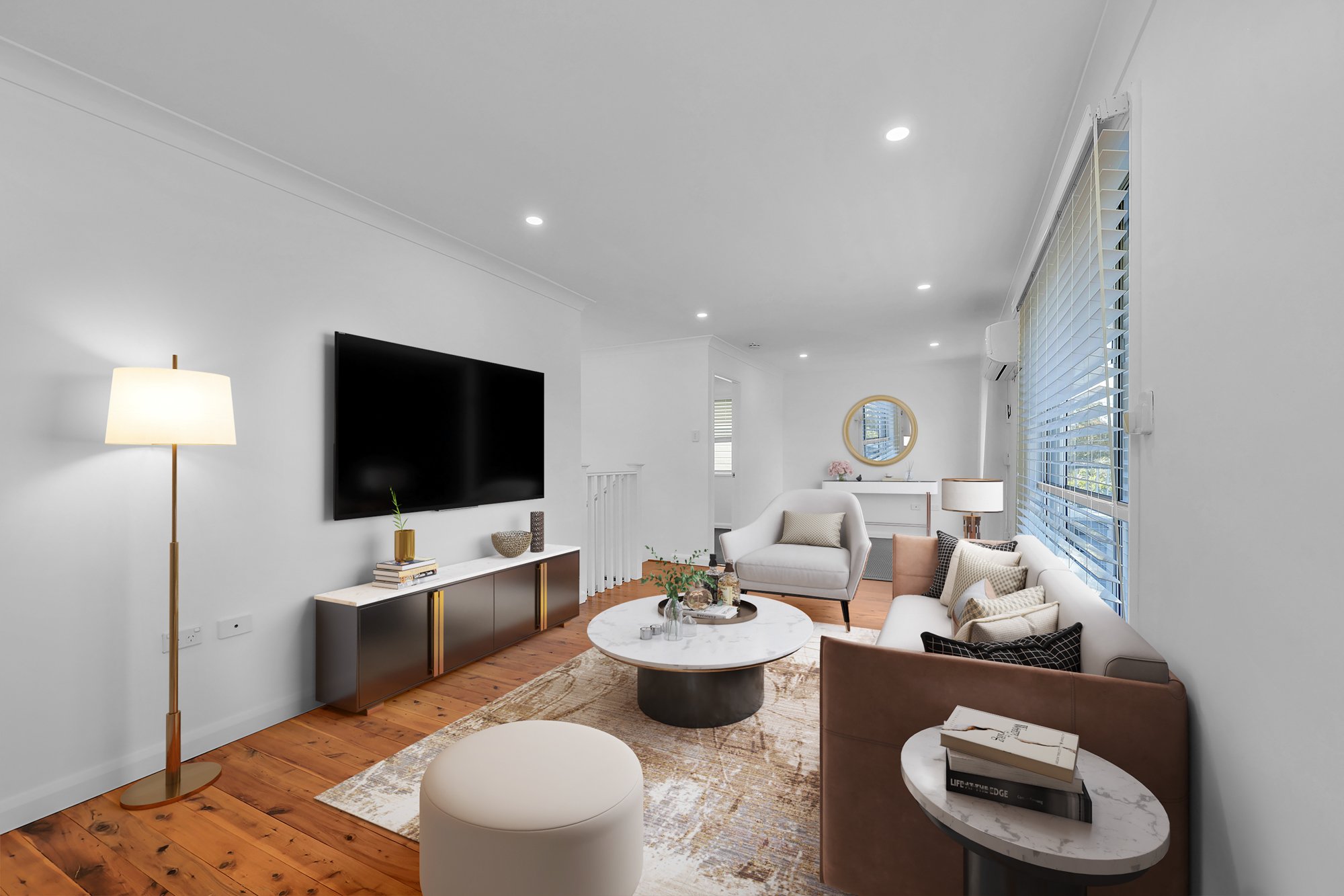 Modern living room with white walls, hardwood floors, a mounted black flat-screen TV, a white armchair, a beige and brown sofa with multiple cushions, a round marble coffee table with decorative items, a floor lamp, a side table with books, a wall mirror, and large windows with white blinds.