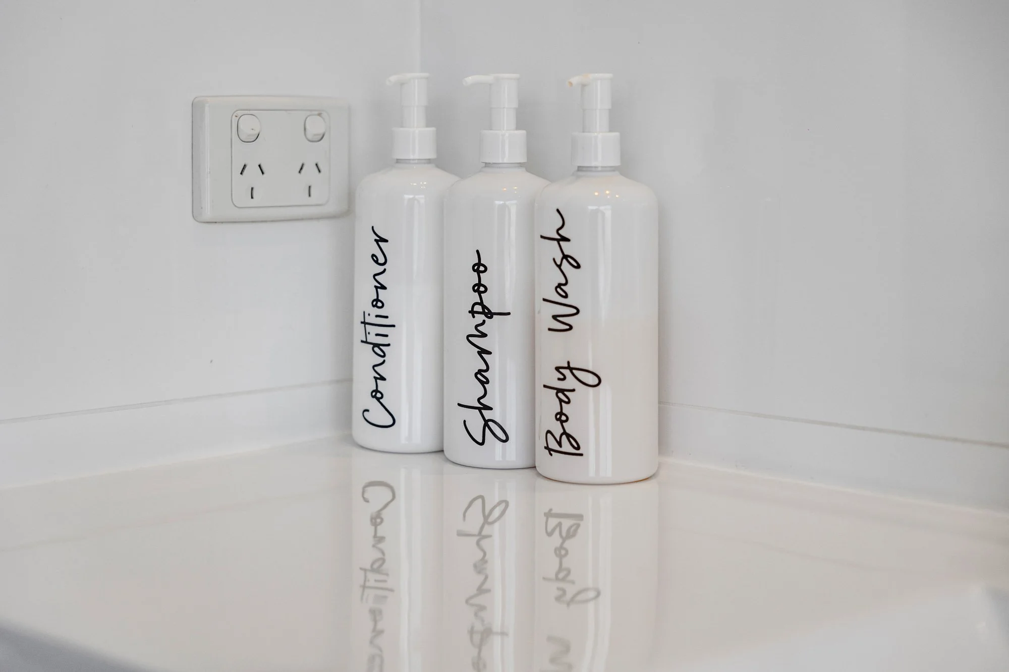 Three white bottles with pump dispensers labeled 'Covenitier', 'Shampoo', and 'Body Wash' in black handwritten script, placed on a white surface near an electrical outlet.