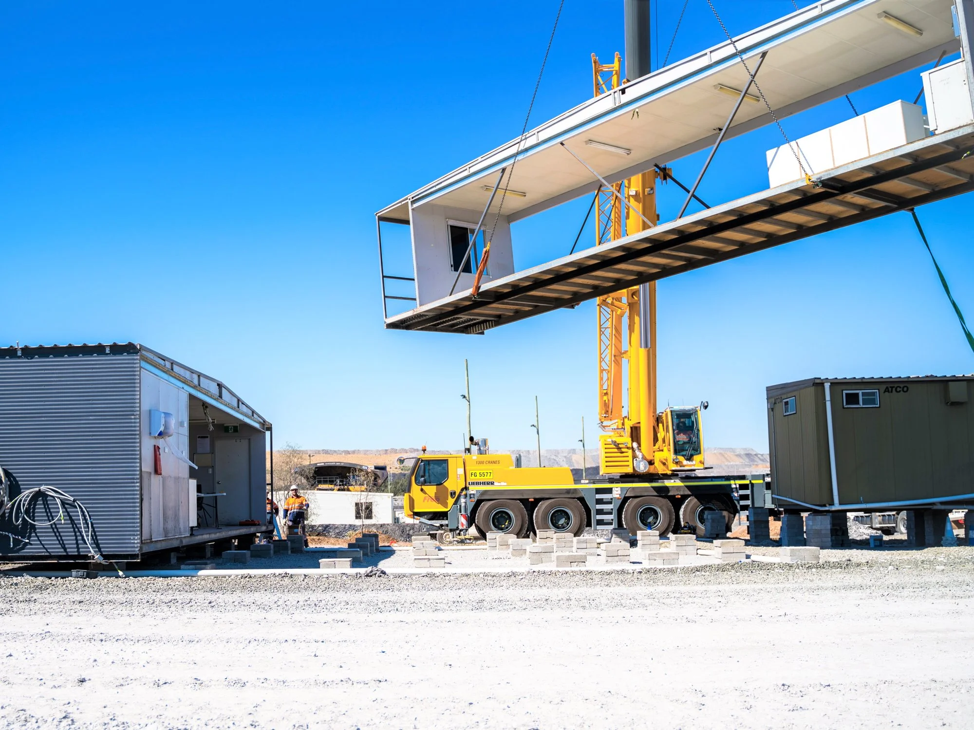 A construction site with a large yellow crane lifting a white modular building. Several temporary structures and workers are visible, with a clear blue sky in the background.