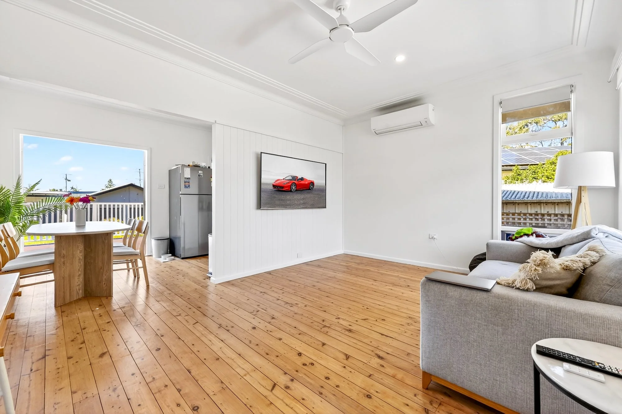 Living room with hardwood floor, white walls, ceiling fan, wall-mounted TV, gray sofa with pillows, side table with remote, floor lamp, window with shades, and a doorway leading to dining area with round table and chairs, refrigerator, and outdoor view.