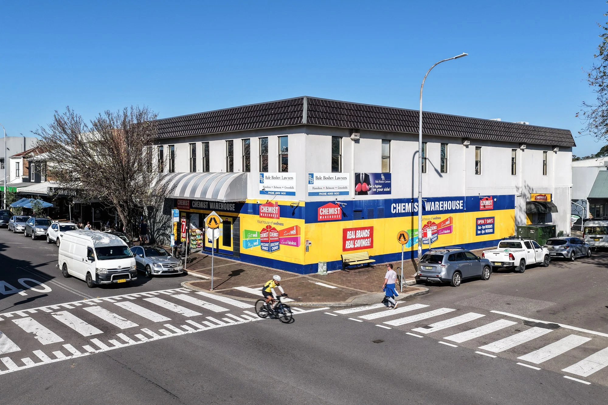 Street corner with parked cars, pedestrian crosswalk, a cyclist, and a storefront with colorful signs advertising chemist and brands in a commercial area.