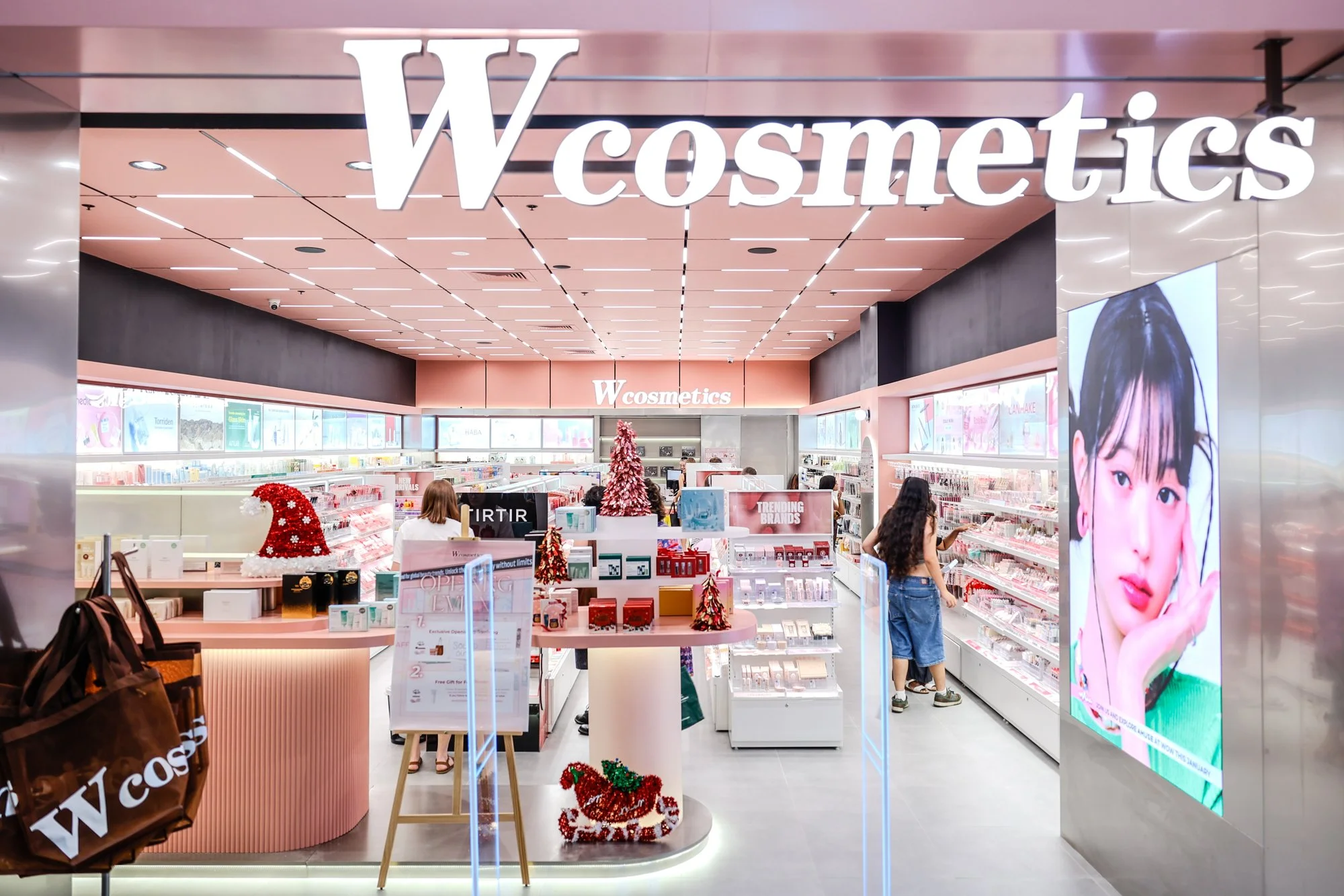 Inside a cosmetics store with pink and white decor, shelves filled with beauty products, a large digital screen displaying a woman's face, and Christmas decorations including a Santa hat and small Christmas trees.